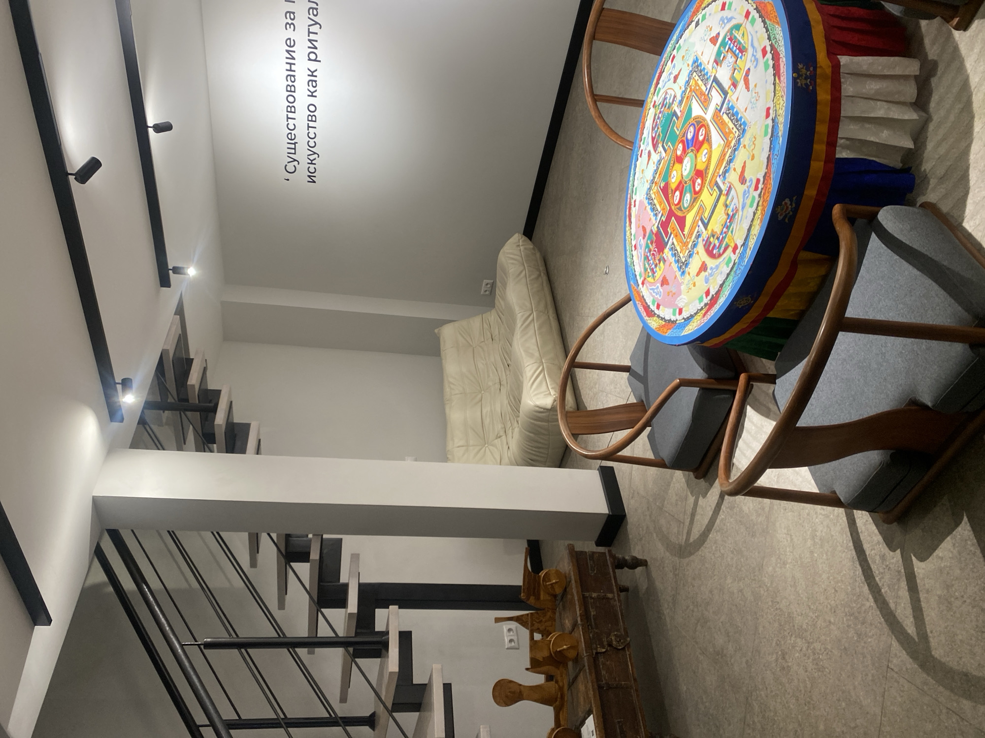 The image shows the interior of a room, possibly a museum or art gallery, located in Moscow, Russia. The foreground features a round table covered with a colorful, mandala-like design. Around the table are several wooden chairs with grey cushions. To the left is a staircase with black metal railings and wooden steps. A light fixture is visible on the ceiling above the staircase. A white, futon-like seating area is situated near the wall. Below the stairs and chairs, a wooden chest-like object rests. The walls are white, and there is text on the wall, likely in Russian. The floor appears to be a light grey tile.
