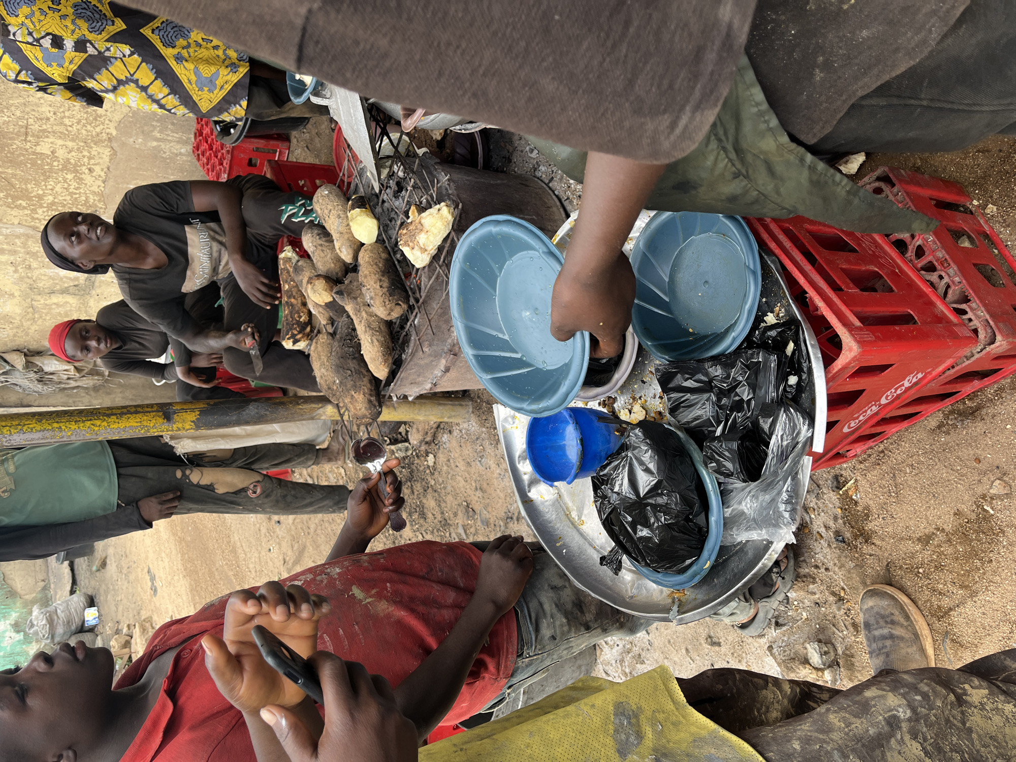 The image depicts an outdoor scene in a dusty environment, likely a market in New Stadium, Nigeria. In the foreground, a young person wearing a red shirt and dirty grey pants holds a black smartphone and a serving ladle. Their left hand is extended, holding the phone. A large metal platter is visible below, containing black trash bags, a blue bucket, and some food scraps. Two blue bowls are also present, one held by another individual whose face is not fully visible. In the background, several people are seated, with one woman wearing a red headscarf and another in a black t-shirt and headscarf. They are positioned near a charcoal grill where several roasted root vegetables are being cooked. A bright yellow and black patterned fabric is draped over someone on the far right. The ground is covered in sand and debris, with red plastic crates visible to the right. The overall impression is a candid, informal street food vending or preparation setting.