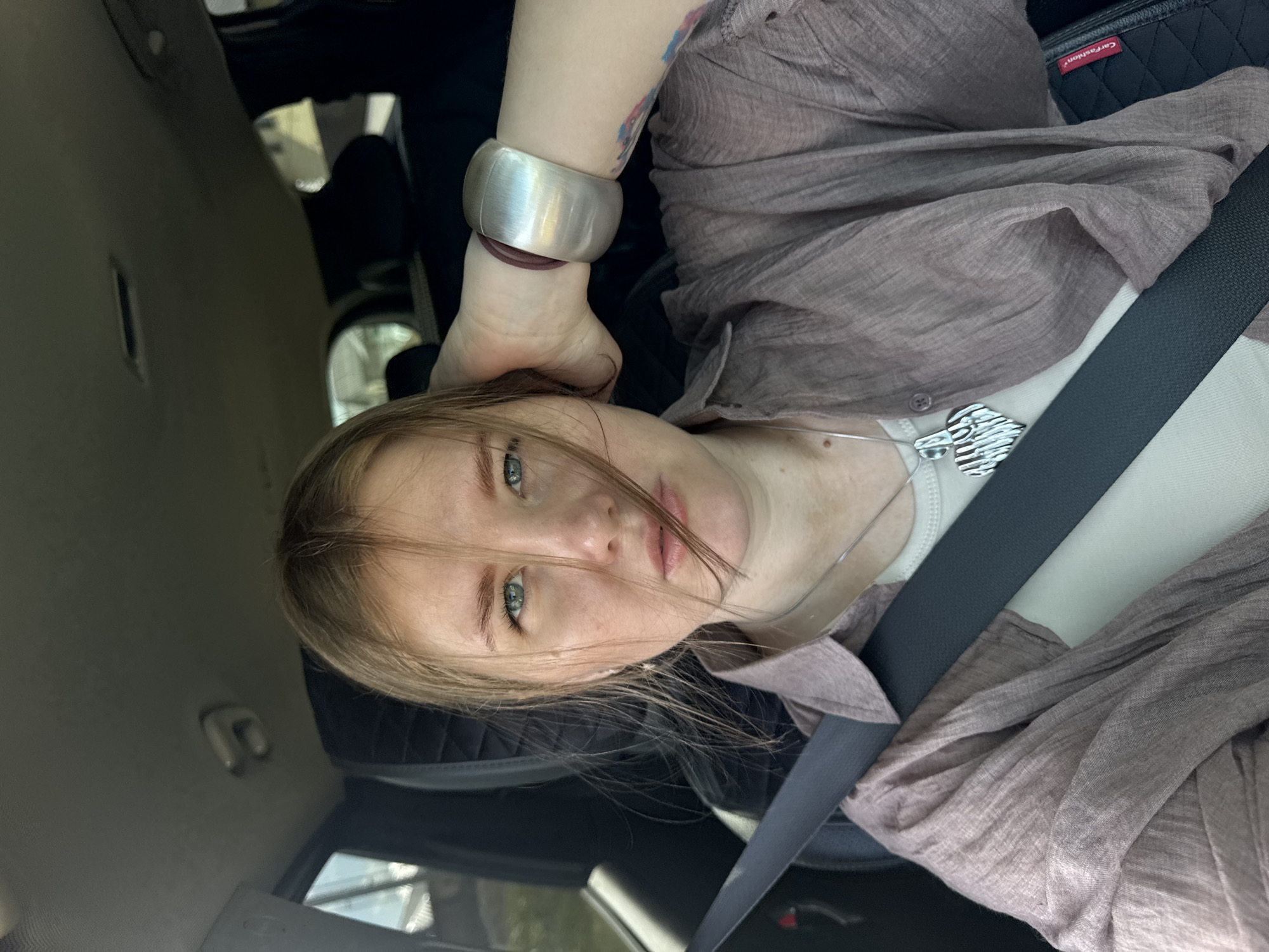 A woman is inside a car, wearing a brown shirt, white undershirt, seatbelt, and a silver bracelet on her left wrist. She is looking at the camera. The car's interior is visible. The location is inside a car, city is not provided.