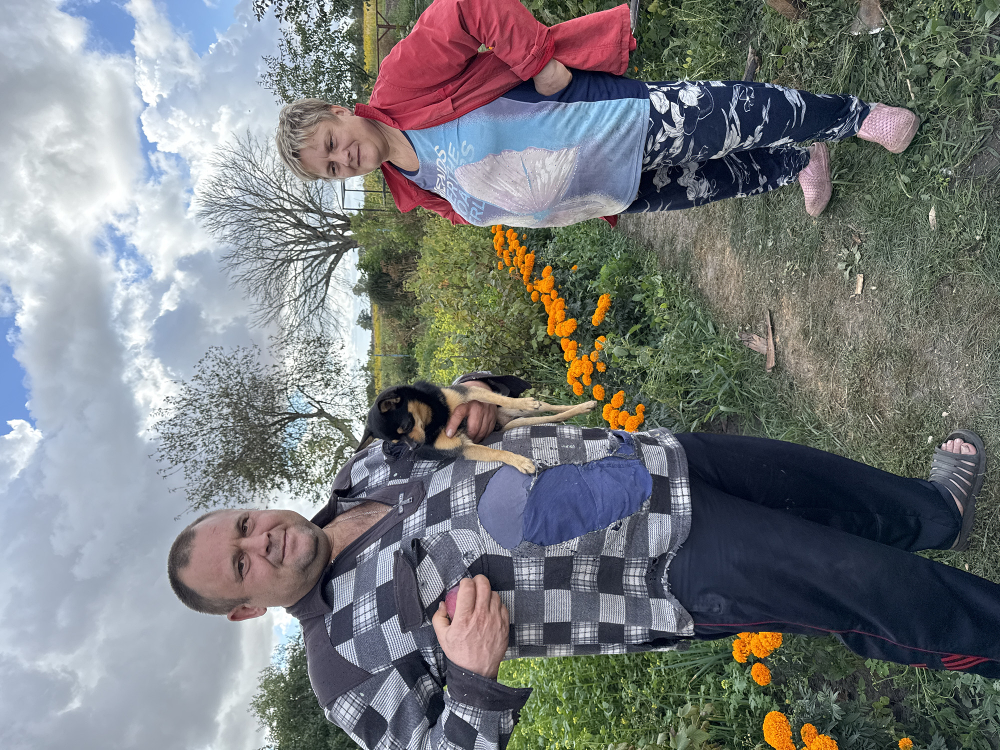 In Fominka, Russia, a man and a woman stand in a garden under a cloudy sky. The man, on the left, holds a small black and tan dog and an apple. He wears a checkered long-sleeved shirt, dark pants, and sandals. The woman, on the right, has blonde hair and wears a red jacket over a blue t-shirt and patterned pants, with pink slip-on shoes. The garden features a path lined with vibrant orange marigolds. Trees and a field of yellow flowers are visible in the background.