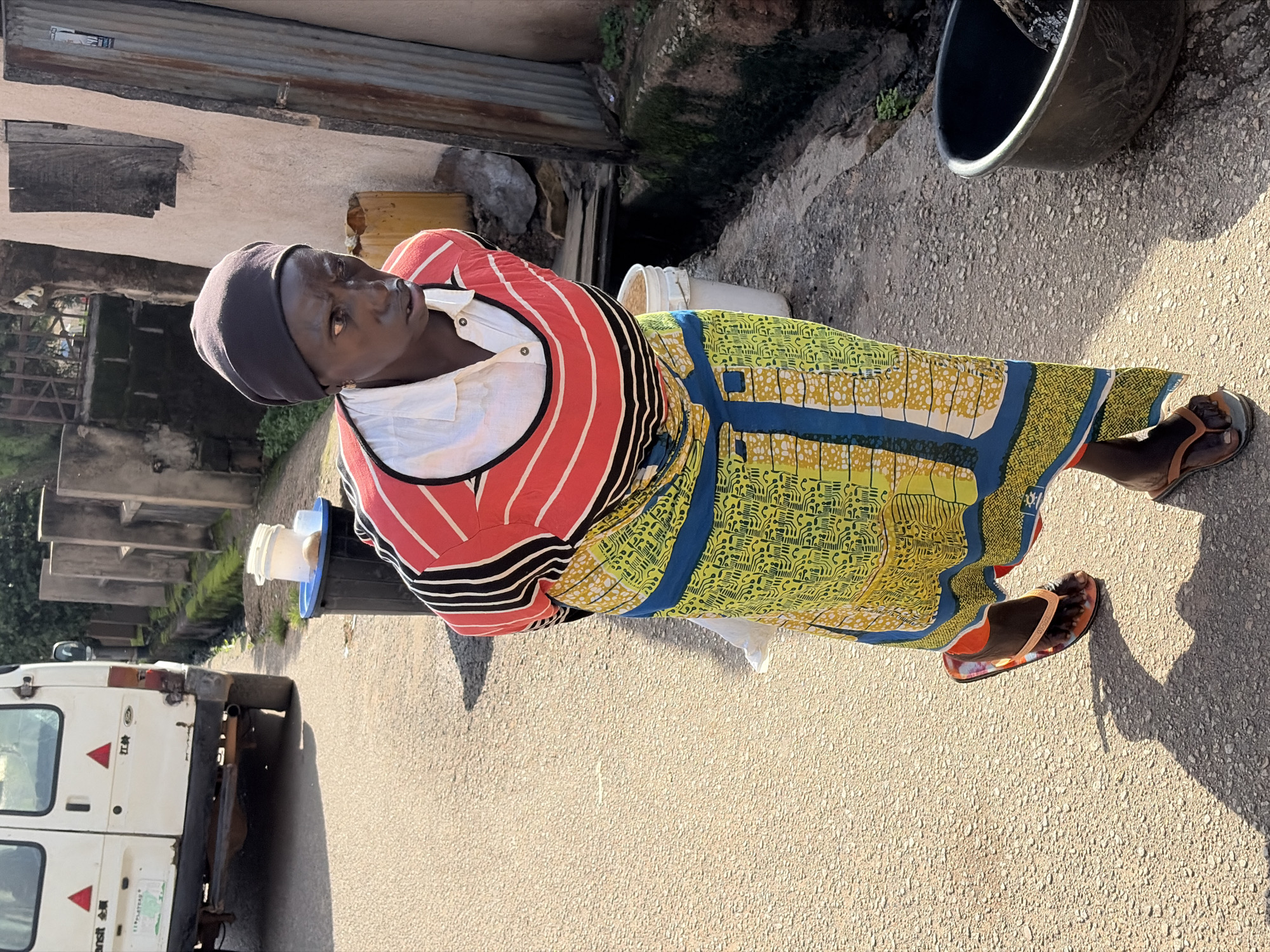 In Jos, Nigeria, a woman wearing a black headscarf, a red and black striped shirt over a white collared blouse, and a vibrantly patterned yellow and blue skirt walks along a paved road. She is wearing orange flip-flops. To her left, a white van is parked. To her right, a black trash bin and a white bucket are visible, along with a large black basin in the foreground. The environment appears to be an urban street with a narrow drainage ditch running alongside a textured wall.