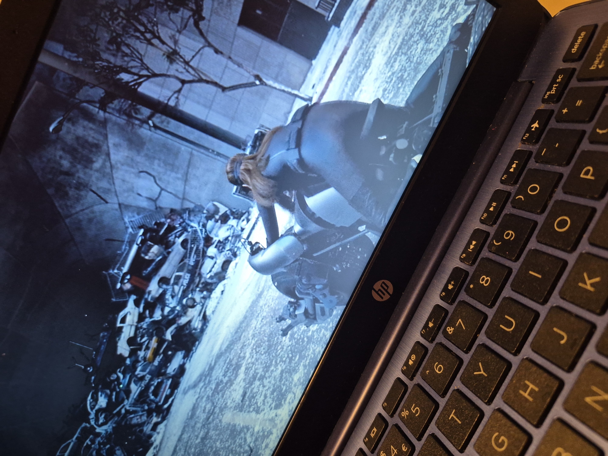 The image displays a laptop screen showing a video game scene. In the foreground, a person is riding a motorcycle, facing away from the camera. The game environment appears to be an urban setting with a junkyard of cars in the background. The laptop itself is visible in the bottom part of the image, with a close-up of its keyboard. The screen shows a blue and gray color palette, suggesting a possibly cold or nighttime setting in the game. There are no specific details in the image that can be used to determine the location in Sandwell, United Kingdom.