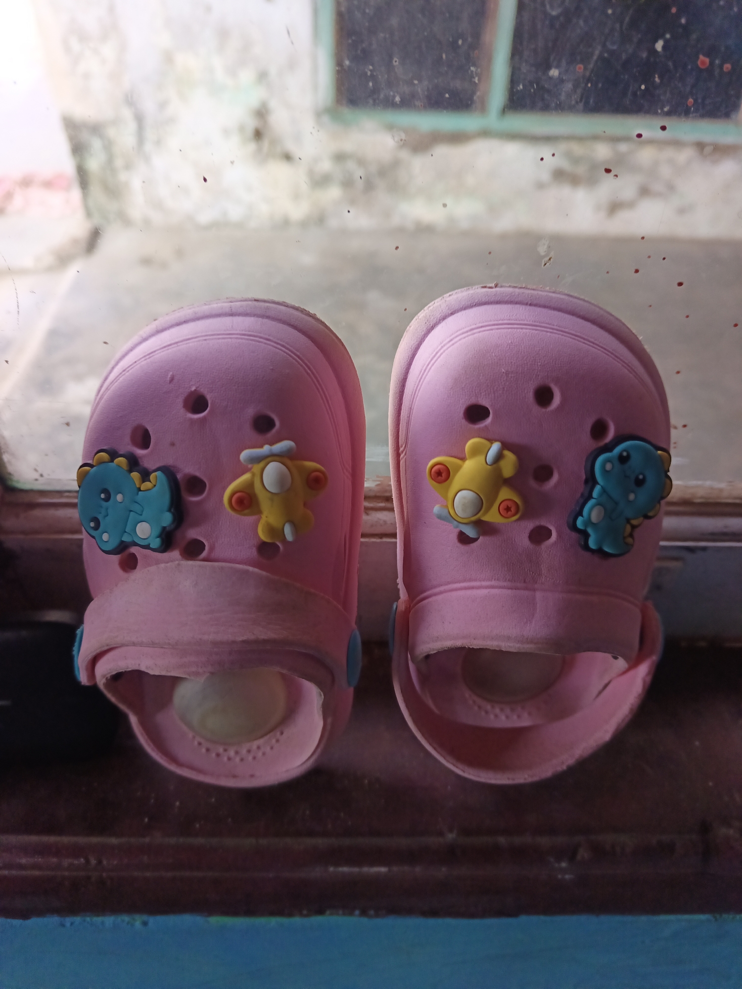 A pair of pink children's clogs with cartoon dinosaur and airplane charms are placed on a dark, wooden surface, likely a windowsill. The clogs are positioned side-by-side. The background shows a blurry view through a dirty window, with hints of a building exterior and possibly a sky. The scene is static and appears to be an indoor shot with natural light. The location is specified as Terbanggi Marga, Indonesia.