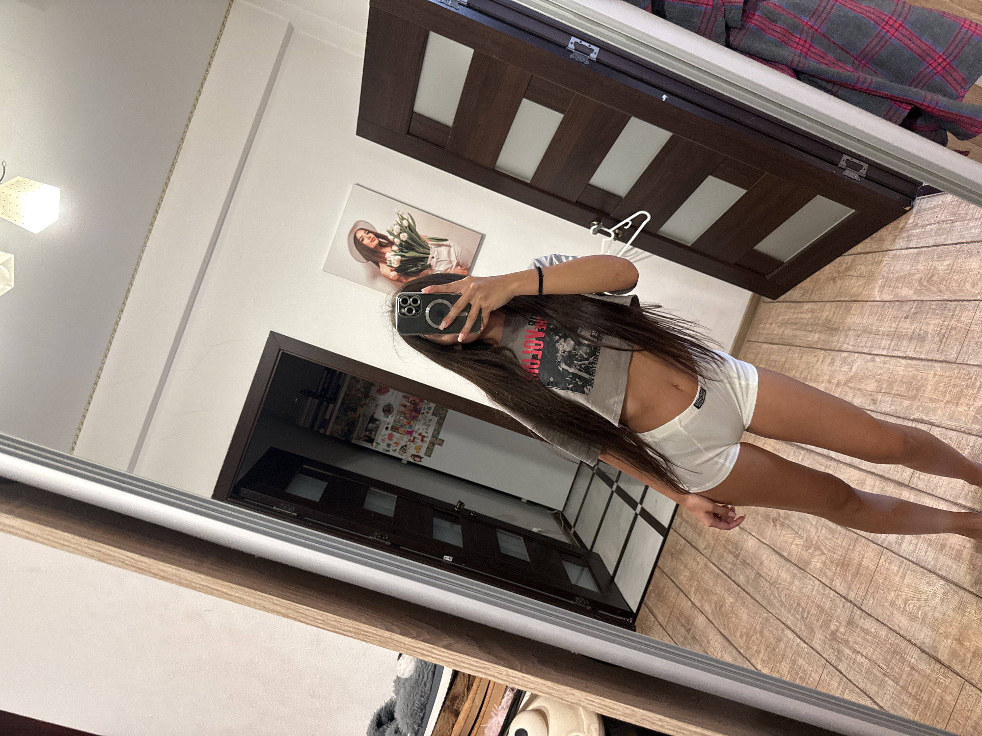 A young woman with long, dark hair stands in a room and takes a mirror selfie. She is wearing a grey cropped t-shirt and white boxer-style shorts. In the reflection, a doorway with a dark wood door and frosted glass panels is visible. On the wall opposite the door, a framed picture of a woman wearing a hat and holding tulips hangs. To the left of the woman, a large mirror reflects a closet with clothes and a stuffed animal. The room has light wood flooring and white walls. The image appears to have been taken indoors, likely in a residential setting.