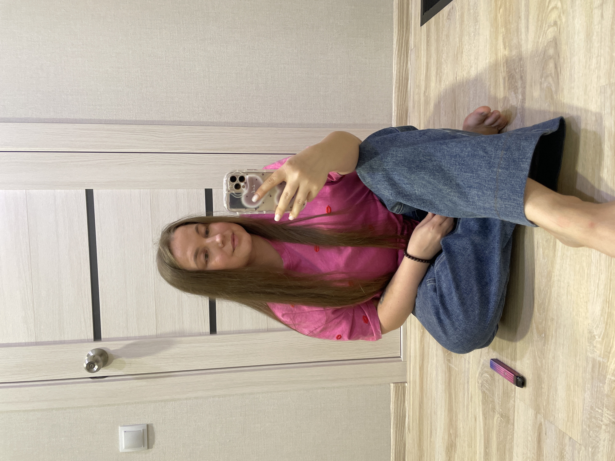 An individual is depicted taking a mirror selfie in an indoor residential setting in Pervomaysky, Russia. A female with long, light brown hair, wearing a pink t-shirt patterned with red lips and wide-leg blue jeans, sits barefoot on a light wood-effect laminate floor. She holds a silver-colored smartphone with three camera lenses in a clear, heart-patterned case with her right hand, angled towards a reflective surface not fully visible. Her fingernails are painted white, and a piercing is visible on her left nostril. A dark beaded bracelet is on her left wrist. To her left on the floor, a gradient pink-to-purple vape pen is positioned. The background features a light-colored wall with subtle texture, a white rectangular light switch, and a light-colored door with two horizontal dark stripes and a silver door knob.