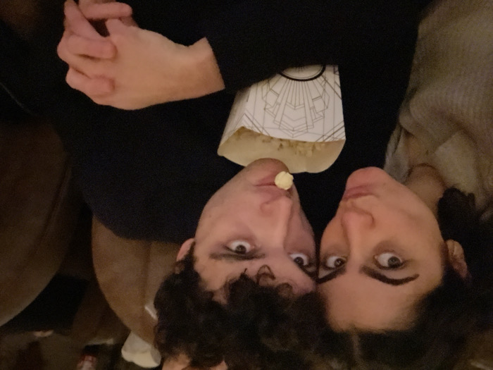 A couple reclines on a brown couch, sharing popcorn. The man has curly dark hair, a popcorn kernel perched on his lip, and wide eyes. Beside him, the woman gazes into the camera with dark curly hair. A bag of popcorn with an art deco design sits between them. Their hands are clasped at the top of the image. Both faces are tilted toward the camera from a low angle.