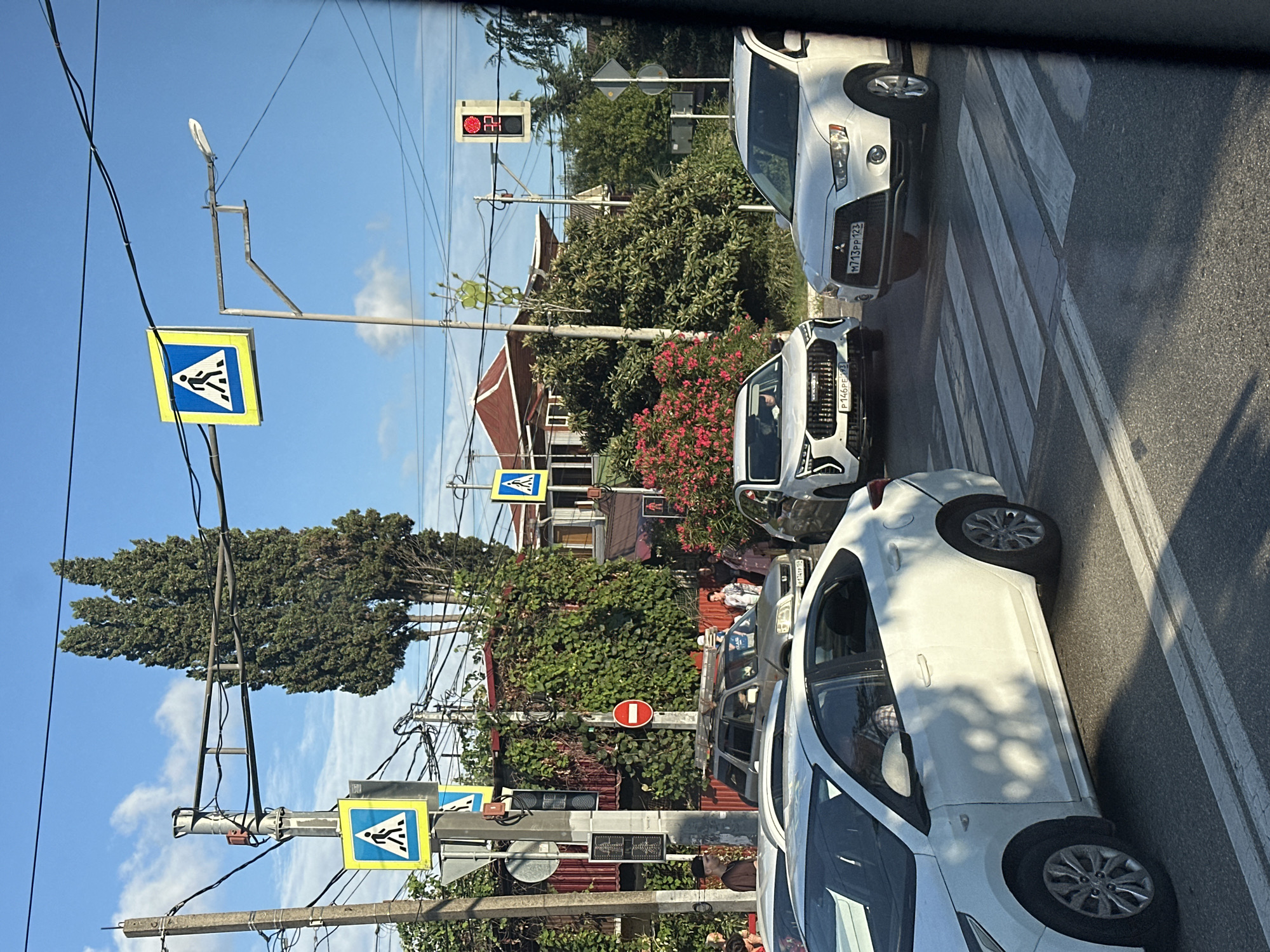 A street scene in Sochi, Russia, depicts traffic at a pedestrian crossing. Multiple cars, including white sedans and SUVs, are stopped at the intersection. Pedestrian crossing signs are visible, accompanied by a traffic light displaying a red signal with a countdown timer showing "32." Overhead wires and streetlights are present, along with lush green trees and foliage. Several people are standing near the intersection, some appearing to be waiting to cross. Buildings with red roofs are visible in the background.