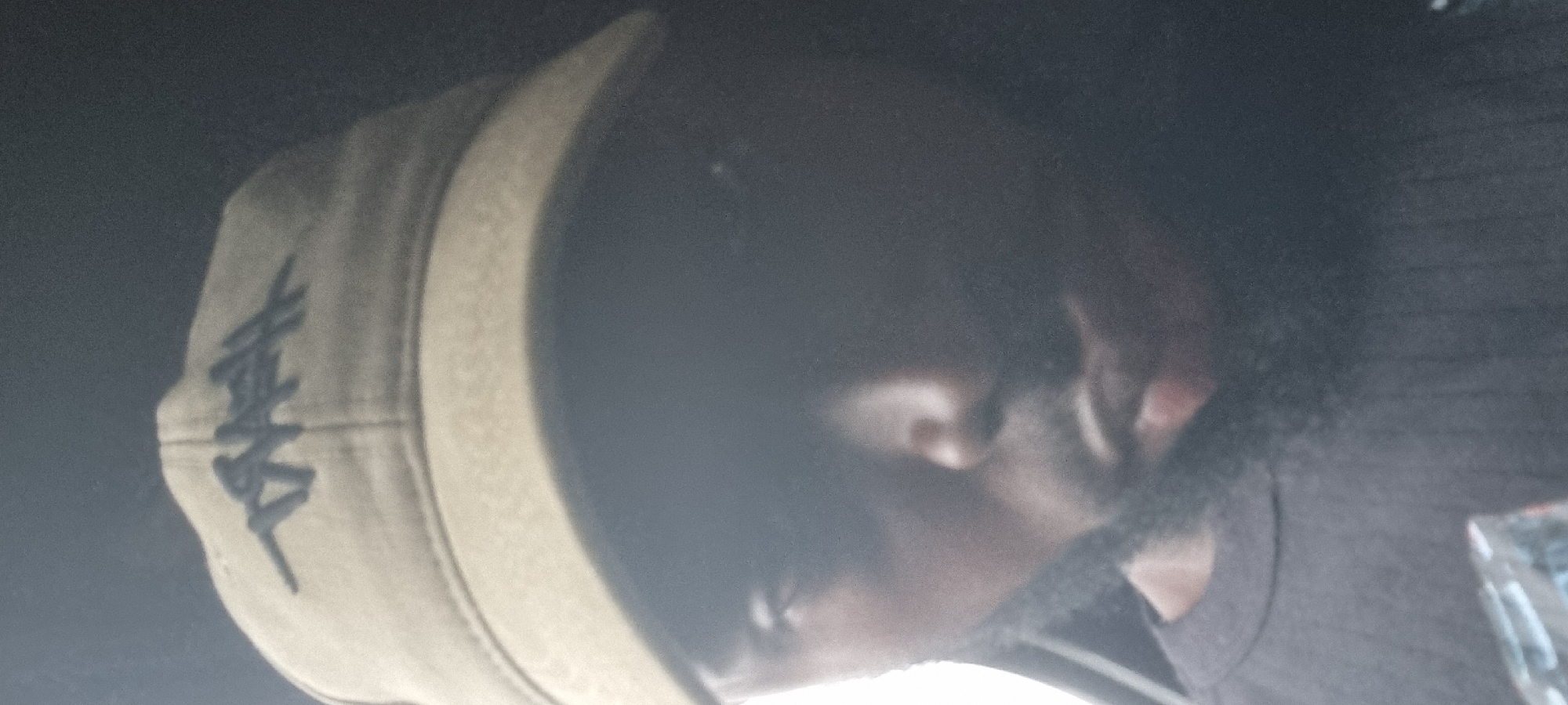 The image shows a close-up, selfie-style photograph of a person wearing a tan baseball cap with a dark logo. The person appears to have dark skin and is wearing a dark, ribbed t-shirt. The lighting is dim, casting shadows across the face and obscuring some details. The background is dark and out of focus, with a bright, out-of-focus area on the left side suggesting a light source or window. The photograph appears to be taken indoors, possibly in a vehicle, due to the way the light hits the person and the dark, enclosed background. There is no visible indication of the location being Ikeja, Nigeria.