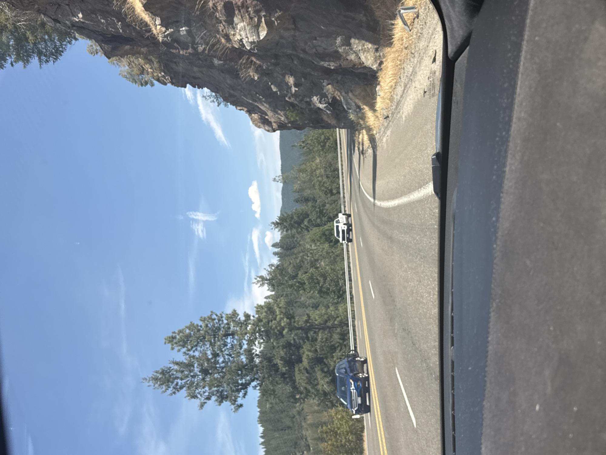 The image shows a view from inside a vehicle, looking out at a winding road. The road has two lanes, with a yellow line marking the center. On the left side of the road, there is a blue SUV driving in the left lane. On the right side of the road, a white pickup truck is driving in the right lane. The road is surrounded by a dense forest of pine trees. To the right of the road, there is a steep, rocky cliff face with some dry grass at its base. The sky is blue with a few wispy white clouds. The scene appears to be in a mountainous or hilly region. The image does not contain any identifiable landmarks or features that would allow for precise location within the city of Pacific, United States.