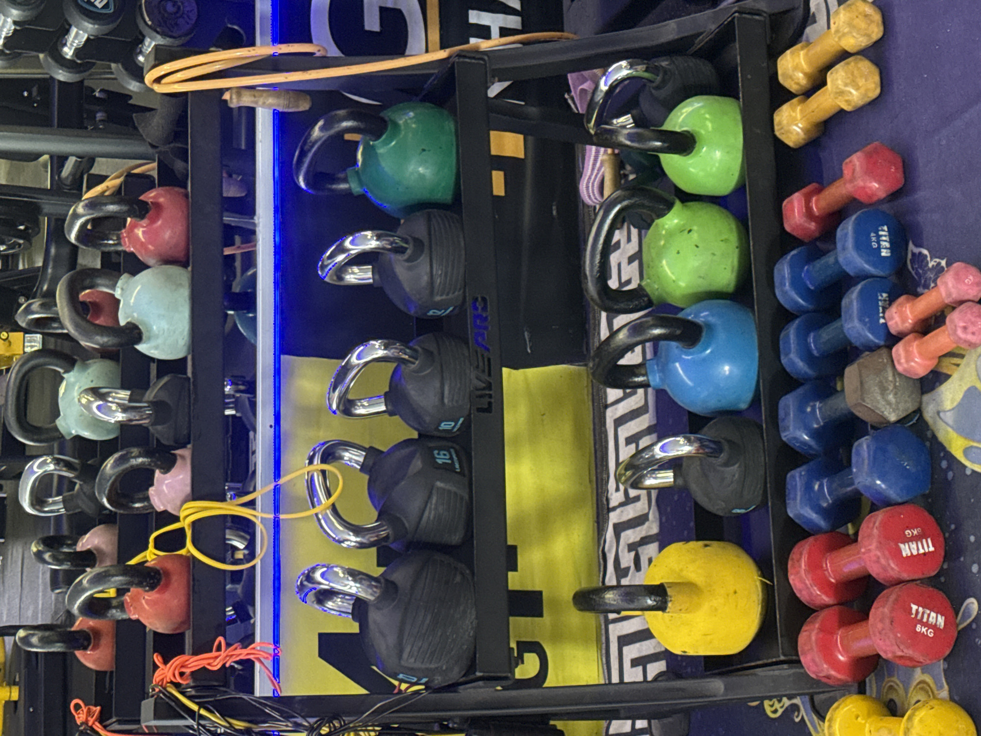 The image shows a weight rack in a gym located in Bac Nha Trang Ward, Vietnam. The rack holds various pieces of weightlifting equipment, including kettlebells and dumbbells.
On the top shelf, there are several kettlebells of different colors: red, pink, light blue, and green. The kettlebells have black handles.
The second shelf from the top contains several black kettlebells, two of which are clearly marked with weights "10" and "16." There is also a green kettlebell on this shelf. The weights are placed on a black rack with metal supports. Behind the weights, there are blurred yellow and white signs with large letters, possibly indicating the gym's name or affiliation. A blue LED light strip runs horizontally below this shelf.
The third shelf from the top displays more kettlebells. A yellow kettlebell is on the left, followed by a black one, a blue one, a green one, and another black one on the right.
The bottom of the image shows a collection of dumbbells. There are red dumbbells, some labeled "TITAN 8KG" and "TITAN 5KG." Blue dumbbells are also visible, as well as yellow dumbbells. Some of the dumbbells are hexagonal in shape.
The overall environment appears to be a gym with a focus on free weights. The lighting is somewhat dim, with the blue LED strip providing a distinct illumination. The floor is covered in a patterned blue matting. The image captures a static display of exercise equipment, with no people present.