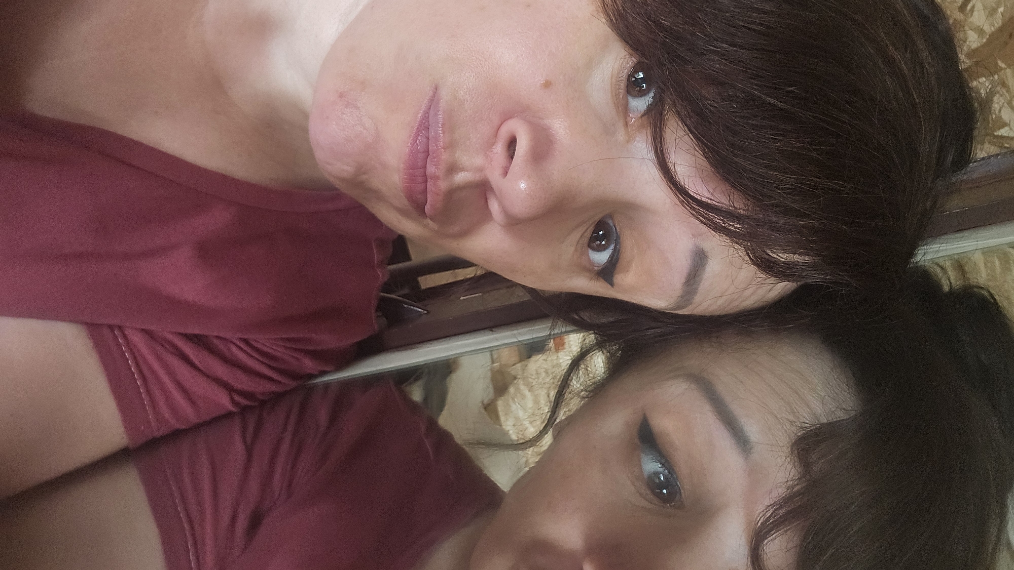 This is a close-up, slightly high-angle, selfie-style shot of a woman with dark brown hair and fair skin. She is wearing a maroon t-shirt. Her left eye is looking directly at the camera, while her right eye is slightly averted. She has winged eyeliner on both eyes. Her lips are slightly parted and appear to be wearing a pinkish lipstick. A reflection of her head and upper body is visible to the right of the frame. The background consists of a textured, light-colored wall and what appears to be a wooden surface. This image was taken indoors, likely in a residential setting in Ермолаево, Russia.