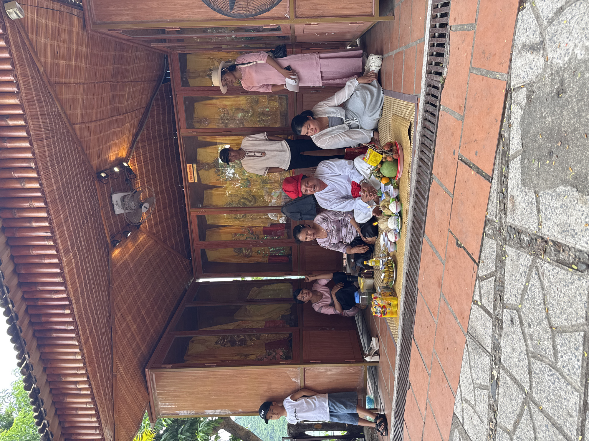 This image captures a scene within an open-air, bamboo-roofed structure that appears to be a cultural or religious site in Nha Trang, Vietnam. In the foreground, several people are seated on woven mats. A central figure, distinguished by a red headdress and white robes, is actively engaged, possibly performing a ritual or ceremony, with a tray of offerings including fruits and what looks like cooked food placed before him. To his right, a woman in light gray attire is also seated, looking towards the offerings.
Behind this central group, a young boy in a white t-shirt and denim shorts stands near a wooden cabinet with glass doors, which display yellow draped fabrics, suggesting religious or ceremonial vestments. To the left of the boy, two women are seated on the matting, one in a pink shirt and black pants, the other in a patterned purple blouse and black pants.
Further back, two individuals stand. A man in a white collared shirt and black pants is looking down, while a woman in a pink dress and a straw hat stands to his right, holding a book or paper. The structure itself is built with wooden framing and walls, with the interior displaying cabinets filled with yellow garments. The roofing is constructed from bamboo poles and woven bamboo mats, and the floor outside is paved with reddish-orange tiles and grey stonework. The overall atmosphere suggests a cultural event or a visit to a place of worship or heritage.