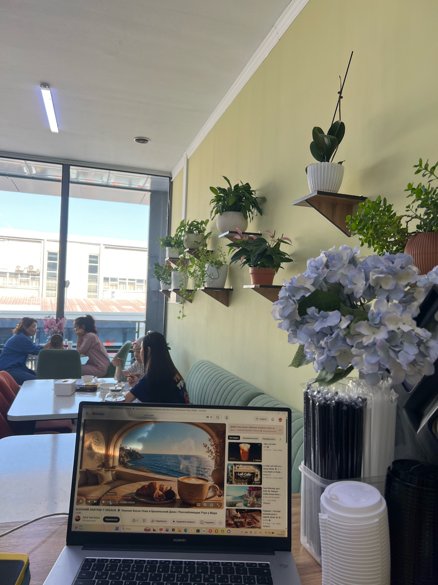 The image shows a cafe interior with people seated at tables. In the foreground, a laptop displays a YouTube video with a scenic ocean view and a cup of coffee. To the right, there are stacks of disposable cups and a container filled with straws. In the background, several individuals are seated at tables, appearing to be engaged in conversation or enjoying their meals. Plants are displayed on shelves along the wall, and large windows offer a view of the outside. The cafe has a modern, relaxed atmosphere. The presence of a "HUAWEI" laptop suggests it is a modern establishment.