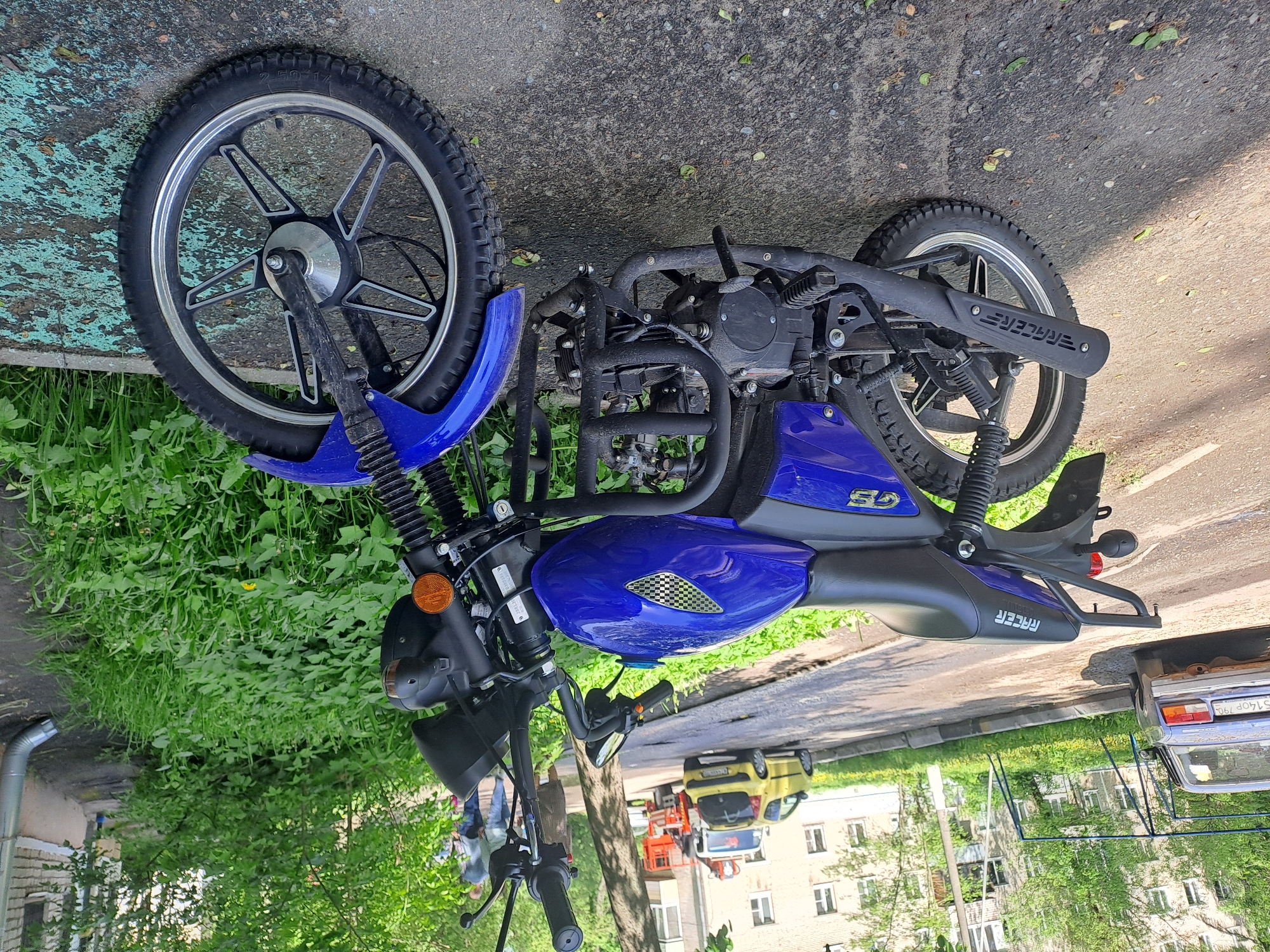 A blue Racer moped rests on the pavement near a grassy area in Bel'kovo, Russia. The moped is parked in front of a silver sedan, with a yellow van and an orange cherry picker visible in the background. A building is situated behind the vehicles. There are a few people blurred in the background.