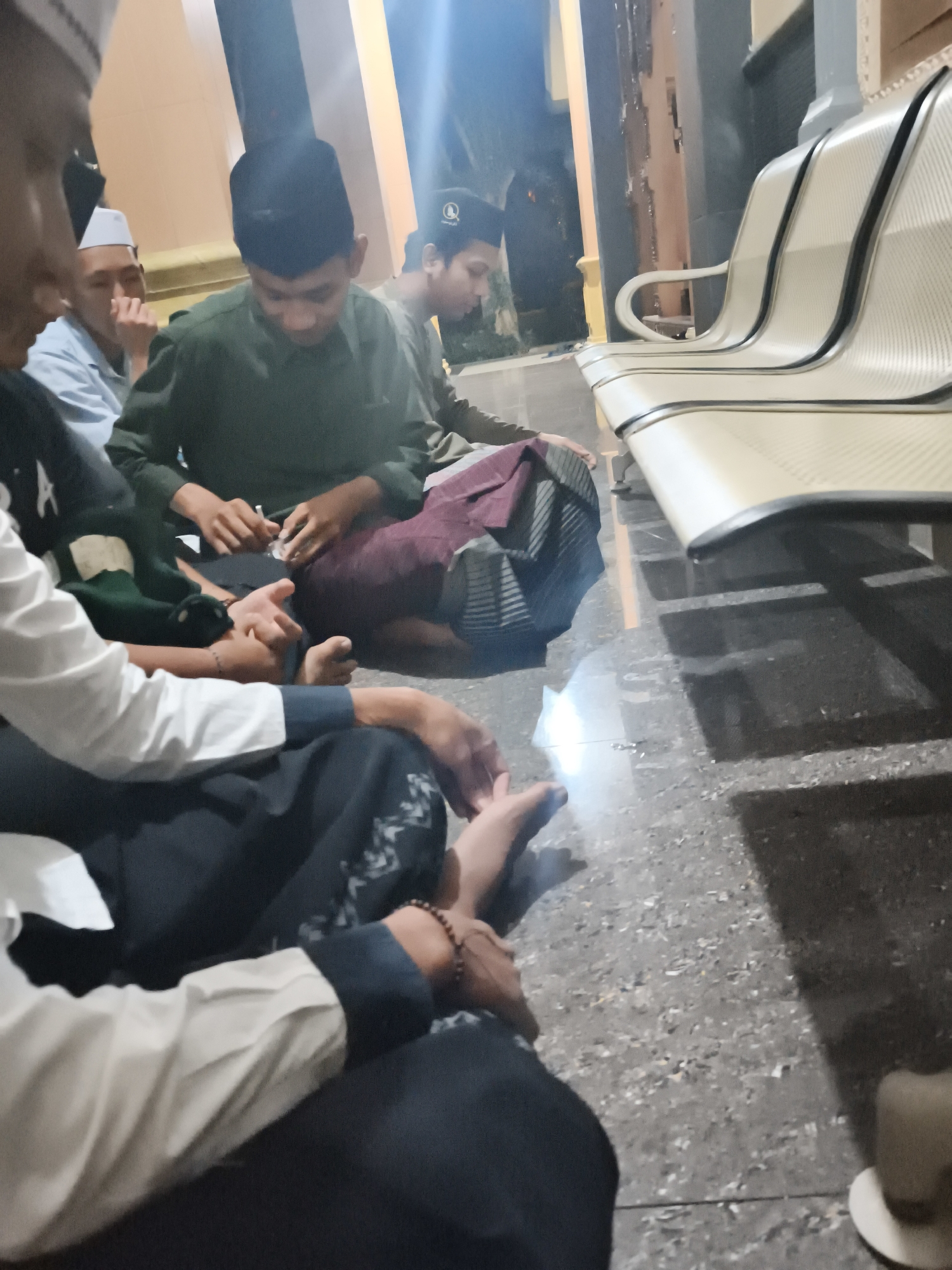 In Ketawang, Indonesia, several individuals are seated on the floor in front of a row of tan, modern-style benches. The scene appears to be indoors, possibly in a waiting area or a place of gathering, with a tiled floor and a light-colored wall in the background. The individuals are dressed in casual attire, with some wearing skullcaps, suggesting a religious or cultural context. One person is holding a piece of fabric, possibly a sarong or a prayer mat. The lighting is dim, with a bright light source casting shadows, indicating it might be evening or an indoor space with artificial lighting. The overall atmosphere is one of quiet contemplation or waiting.