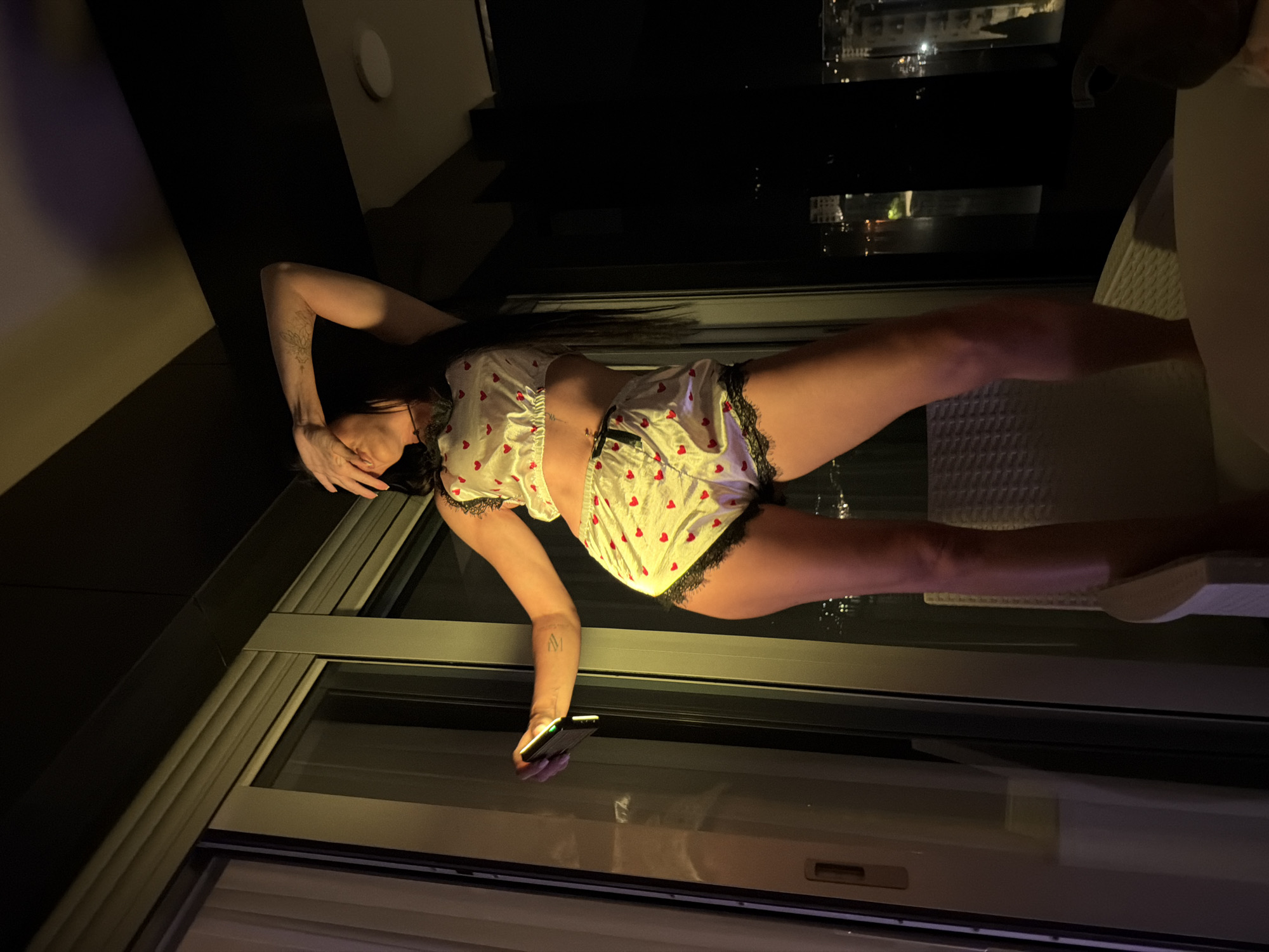 The image depicts a nighttime scene, primarily featuring a young woman in what appears to be a modern indoor setting with a view of an urban or semi-urban landscape. The image is rotated 90 degrees clockwise.
**Subjects:** The central subject is a woman with long, dark hair. She has visible tattoos, including a floral or leafy design on her upper left arm and what appears to be the letters "M" and "V" on her right forearm.
**Objects:** She is dressed in a two-piece pajama set, consisting of a white or cream-colored crop top and matching shorts, both decorated with red heart patterns and black lace trim. In her right hand, held low, she carries a dark-colored smartphone. Part of a light-colored, possibly woven, outdoor chair or piece of furniture is visible on the right side of the frame. Indoors, a white circular light fixture is mounted on a dark wall.
**Scene, Setting, and Activity:** The woman stands beside a large glass sliding door or window, through which distant city lights are visible. The scene is dimly lit, suggesting late evening or night. Her left hand covers part of her face, while her right arm is extended downwards, holding the phone. Her pose appears relaxed or pensive, possibly a self-portrait or taken by another person. Reflections of the exterior city lights, as well as a faint reflection of her lower body, are visible on the glass surface. The setting is likely a balcony or room in a hotel or apartment building, providing a nocturnal city view.
**Notable details:** The time of day is clearly night, indicated by the dark sky and illuminated distant buildings. The interior lighting is subdued. No discernible text is visible beyond the stylized letters of the tattoo on her arm. The location context of Boreti, Montenegro, suggests a coastal urban environment.