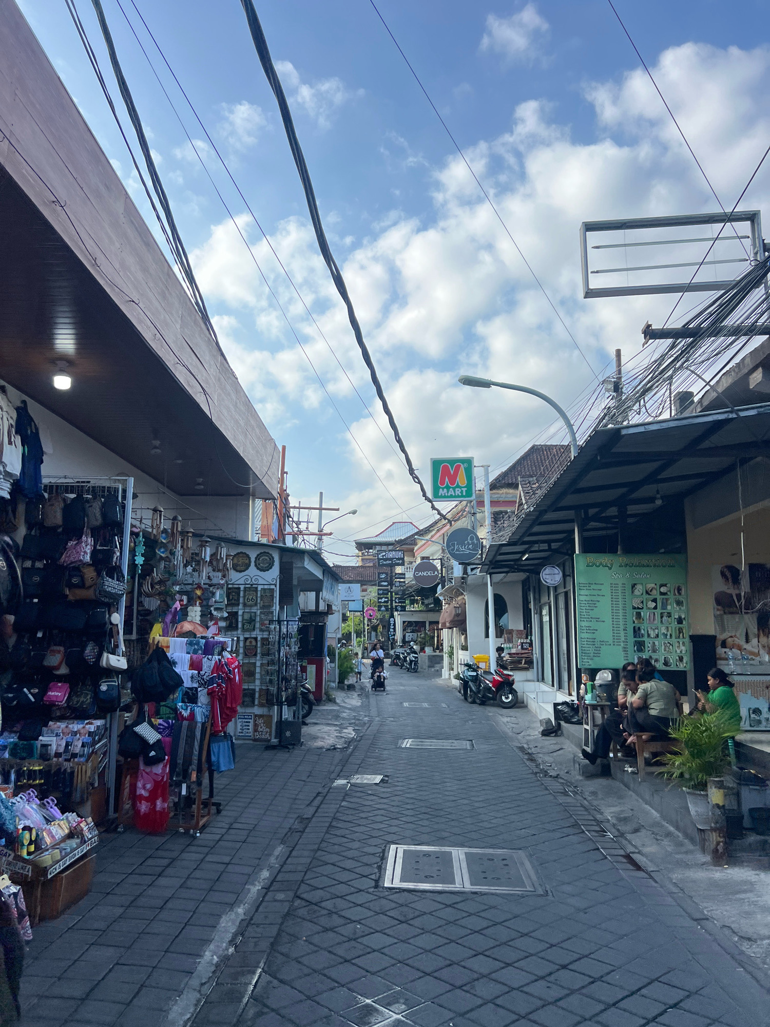 The image depicts a street scene in Kuta, Indonesia, likely during daylight hours. The street is paved with dark grey interlocking bricks and is lined with shops on both sides. The shops on the left display various goods, including handbags, clothing, and souvenirs, with racks and tables set up on the sidewalk. On the right, shops appear to be more service-oriented, with a "Body Relaxation Spa & Salon" sign prominently visible, featuring images of hairstyles and service menus. Several people are seated outside this establishment.
The architecture is typical of a commercial street in Bali, with covered awnings extending from the buildings. Electrical wires are strung across the sky, supported by poles and attached to the buildings, indicating a functional but somewhat untidy urban infrastructure. A street lamp is visible on the right. Further down the street, the scene suggests continued commercial activity with more shops and people. A few motorcycles are parked and one is being ridden in the distance, indicating motorized transport is in use. The sky is partly cloudy, with patches of blue sky visible between white clouds. The overall impression is that of a busy, pedestrian-friendly commercial area.