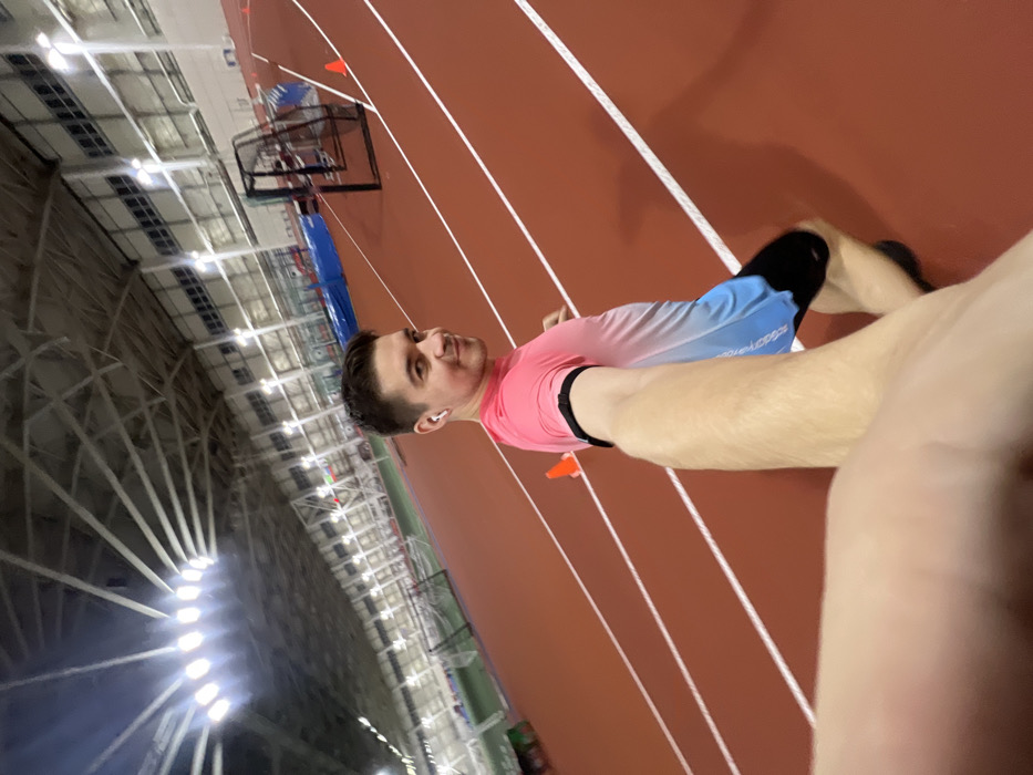 A man takes a selfie at an indoor track. He wears a pink and blue gradient long-sleeve shirt, black running shorts, and white wireless earbuds. A black fitness tracker is visible on his arm. The track is red with white lane markers. An indoor sports complex with a metal frame and bright overhead lights is in the background. There is a basketball hoop near the track. Orange cones are placed on the track.