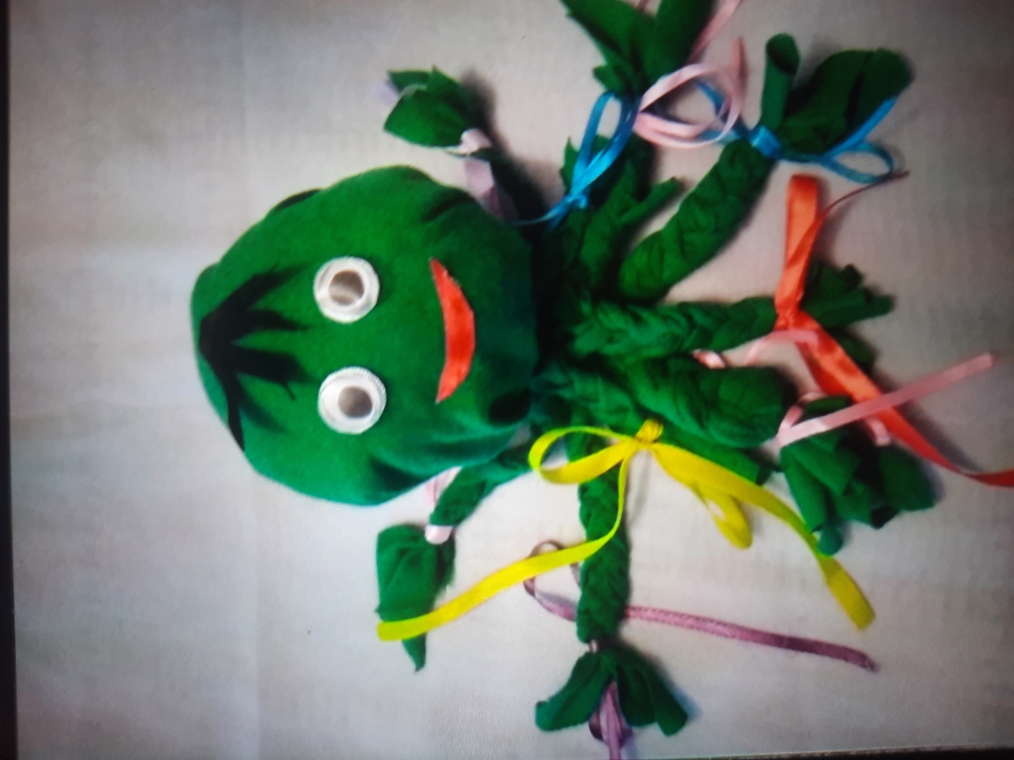A handmade green fabric octopus stuffed toy is displayed on a light-colored, textured surface. The toy has a round head with two white googly eyes and a red fabric mouth. Black fabric pieces are attached to the top of the head, resembling hair. The octopus has eight fabric tentacles, some of which are braided. Various colored ribbons, including yellow, pink, blue, and orange, are tied to the tentacles and trail down. The lighting suggests an indoor setting, likely a domestic environment. There is no indication of the city of Rosenheim, Germany, or any specific location within it.