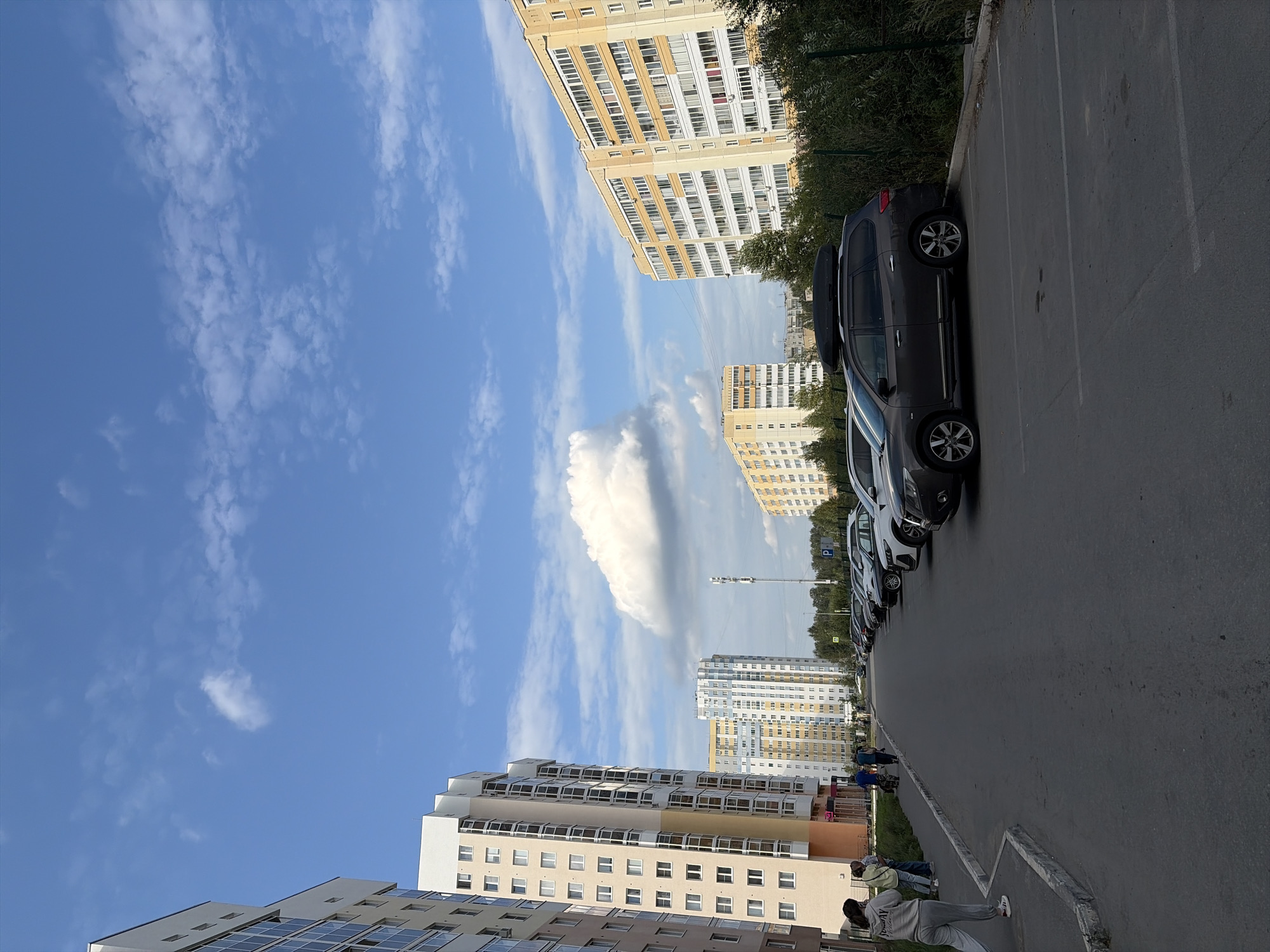 The image depicts a street scene in Yekaterinburg, Russia, likely a residential area, with tall apartment buildings lining one side of a paved road. The sky is blue with scattered white clouds, suggesting daytime. Several cars are parked along the right side of the road. In the foreground, on the left side, are two women walking away from the camera, and further down the sidewalk, several other people are walking. The apartment buildings are modern, featuring multiple floors with many windows. One building in the distance has a telecommunications tower in front of it. The overall environment appears to be a well-maintained urban setting.