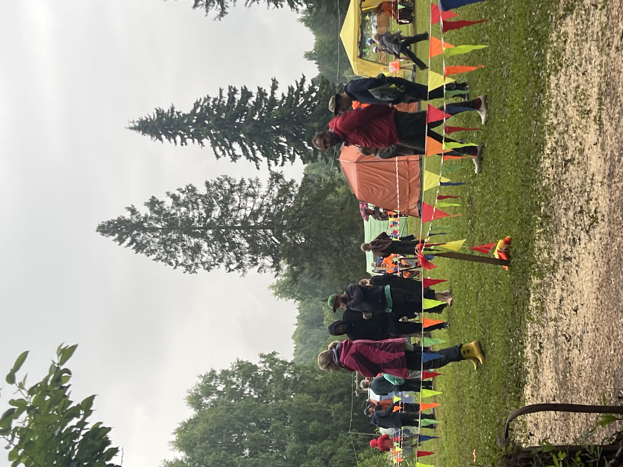 The image depicts an outdoor event, possibly a competition or festival, in a grassy field surrounded by trees, with a cloudy sky. In the foreground, a string of colorful triangular flags stretches across the grass. Several people are visible, some standing and conversing, while others are walking. They are dressed in casual outdoor clothing, with some wearing hooded jackets, suggesting potentially cool or damp weather. Tents, one orange and one yellow, are set up in the background, indicating a temporary setup for the event. The ground appears to be a mix of grass and dirt, with some patches of mud. The overall atmosphere suggests a communal gathering in a natural, rural setting, possibly near the village of Sergino, Russia, given the context.