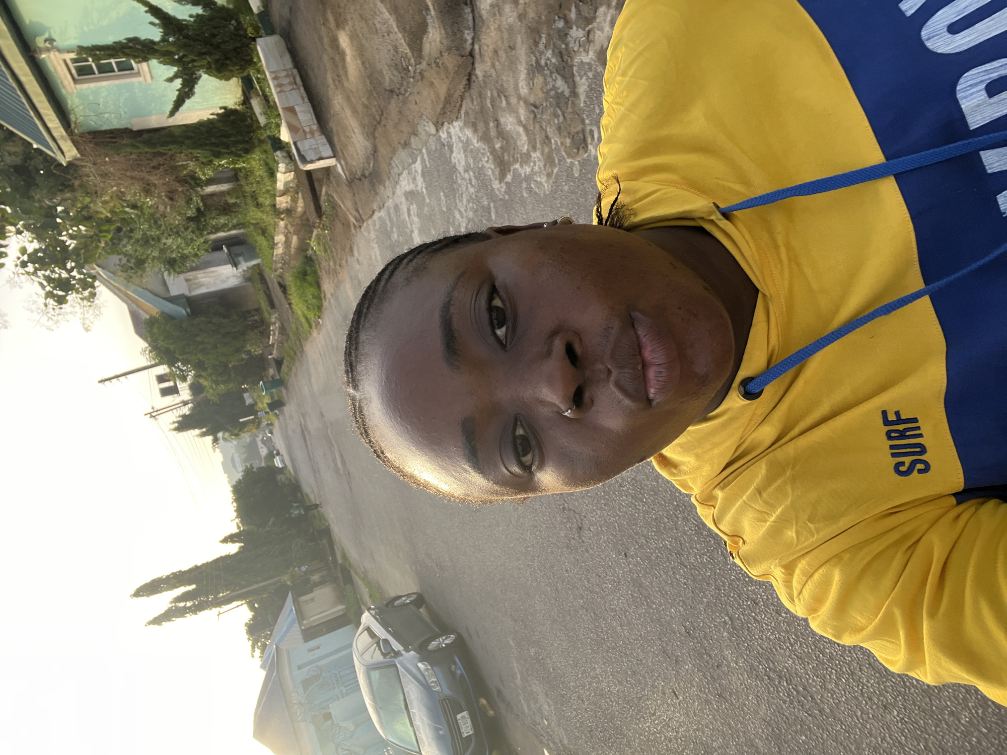 A selfie of a person with dark skin, braided hair, and a nose piercing wearing a yellow and blue hoodie with "SURF" printed on the sleeve. The background reveals a street scene in Abuja, Nigeria, with cars parked along the roadside, houses, trees, and buildings visible under bright sunlight. The road is paved. The image is slightly rotated.
