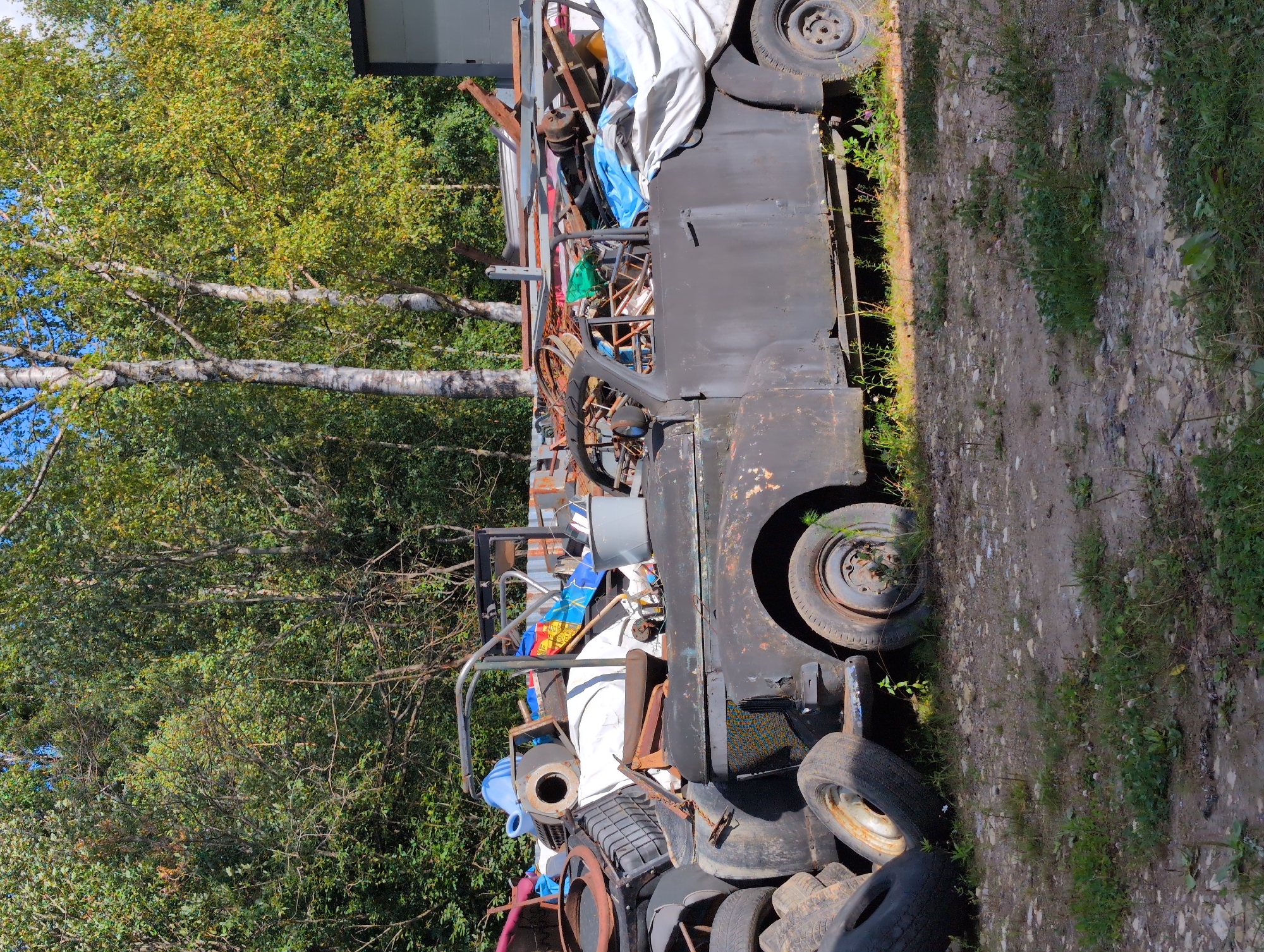 An outdoor scene depicts a damaged, dark-colored vintage car covered in a white tarp on the right side, amidst a collection of tires and assorted debris. The car's front end is visible on the left, and its body is rusted and worn. Stacked tires are piled to the left of the car. In the background, a dense forest of green trees with some yellowing foliage is visible. The ground is covered in dirt and scattered rocks. A partially visible building structure with a gray exterior is present on the far right edge of the frame. The sky is clear and blue. The scene appears to be an outdoor junkyard or storage area, possibly in Novoe Devyatkino, Russia, given the context. There are no people or animals present in the image.