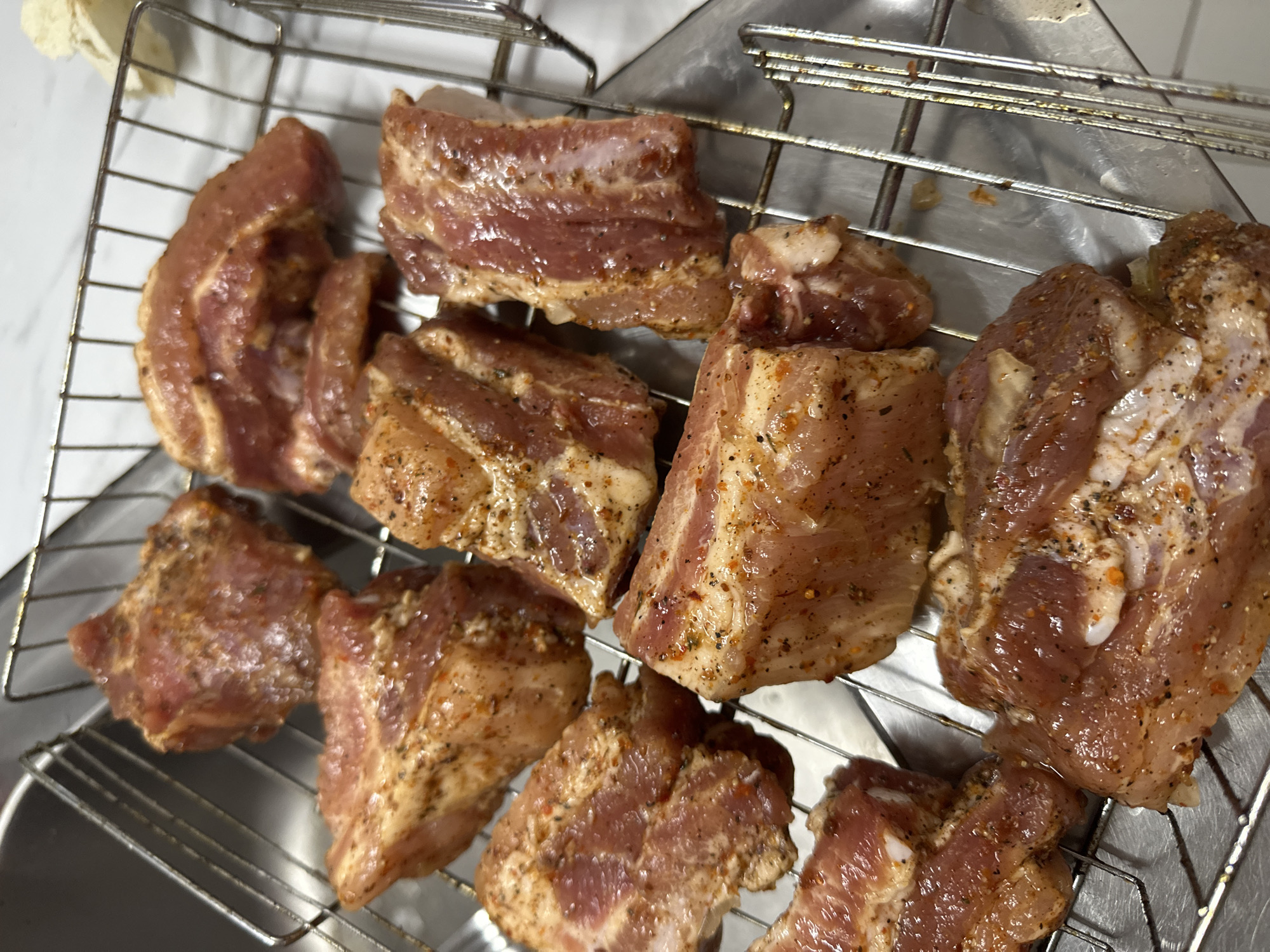The image displays a metal rack holding multiple pieces of raw pork belly, seasoned with spices. The pork belly is cut into rectangular portions and arranged closely together on the rack. The seasoning appears to be a mixture of black pepper, salt, and possibly paprika or chili flakes, giving the meat a reddish-brown hue. The rack itself is made of stainless steel and rests on what appears to be a stainless steel surface, possibly a kitchen counter or sink basin. The background is blurred but suggests a clean, possibly domestic, kitchen environment. There are no people or animals visible in the image. The lighting is bright, highlighting the texture and marbling of the pork. The image was taken from a slightly elevated angle, looking down at the rack of meat. No specific landmarks or features that would indicate the city of Verkhniy Yurt, Russia are present.