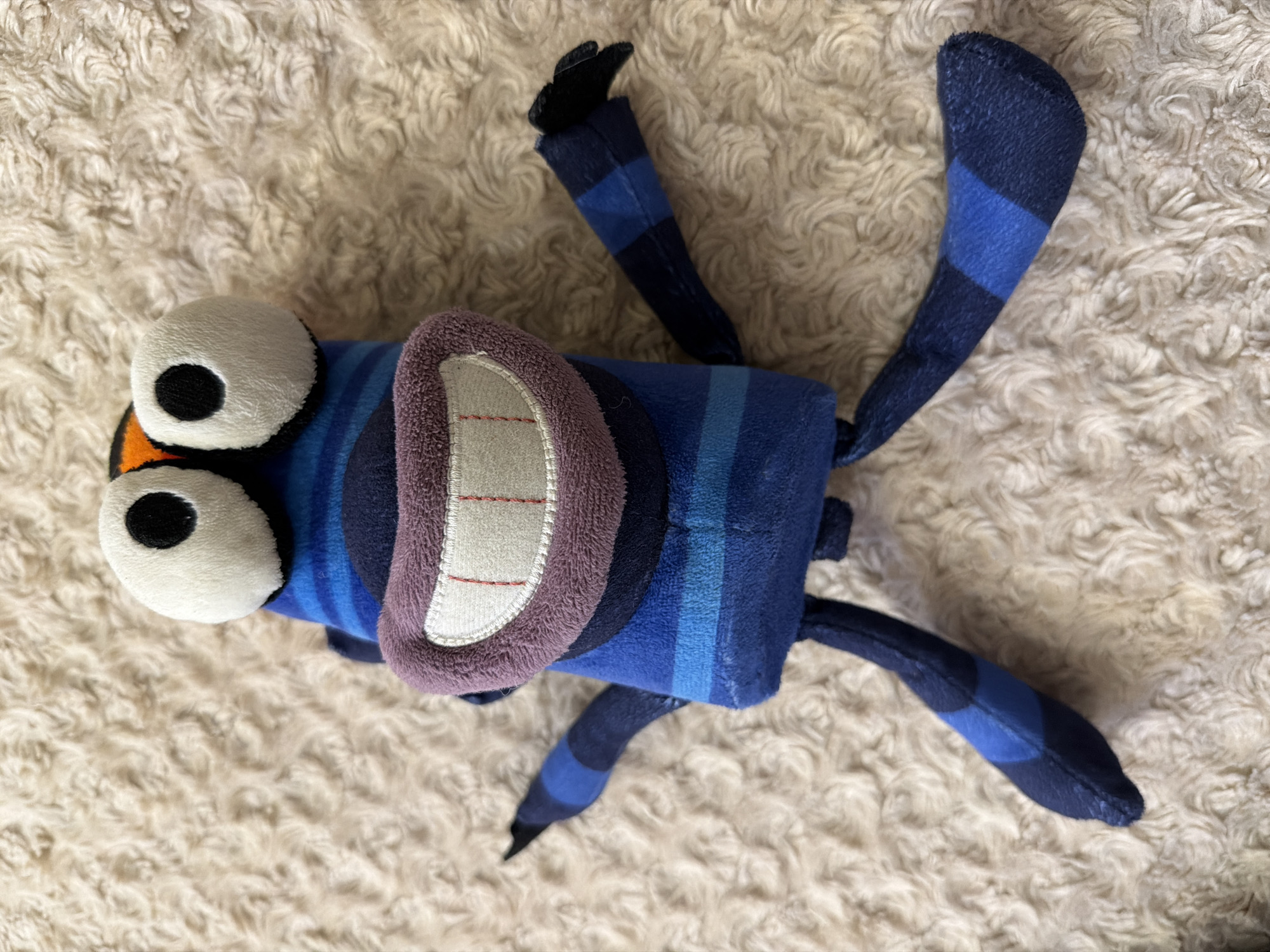 This is a top-down, medium shot of a blue, striped plush toy lying on a fluffy, cream-colored textured surface. The toy is elongated and resembles an insect or a fantastical creature. It has two large, white, round eyes with black pupils, and a prominent mouth with purple lips and white teeth. The toy's body is segmented with alternating dark blue and lighter blue stripes. It has multiple appendages, appearing to be legs or antennae, also striped in blue. One appendage on the upper left has a small black tip. The plush toy is the sole subject, and no activity or event is taking place beyond its static pose. The lighting appears to be indoor, possibly natural light from a window, as there are no harsh shadows. There are no visible text elements in the image. The overall emotion conveyed is playful and whimsical, characteristic of a children's toy. The location context of Krasnoyarsk, Russia, provides a geographical setting but is not directly indicated by visual cues in the image itself.