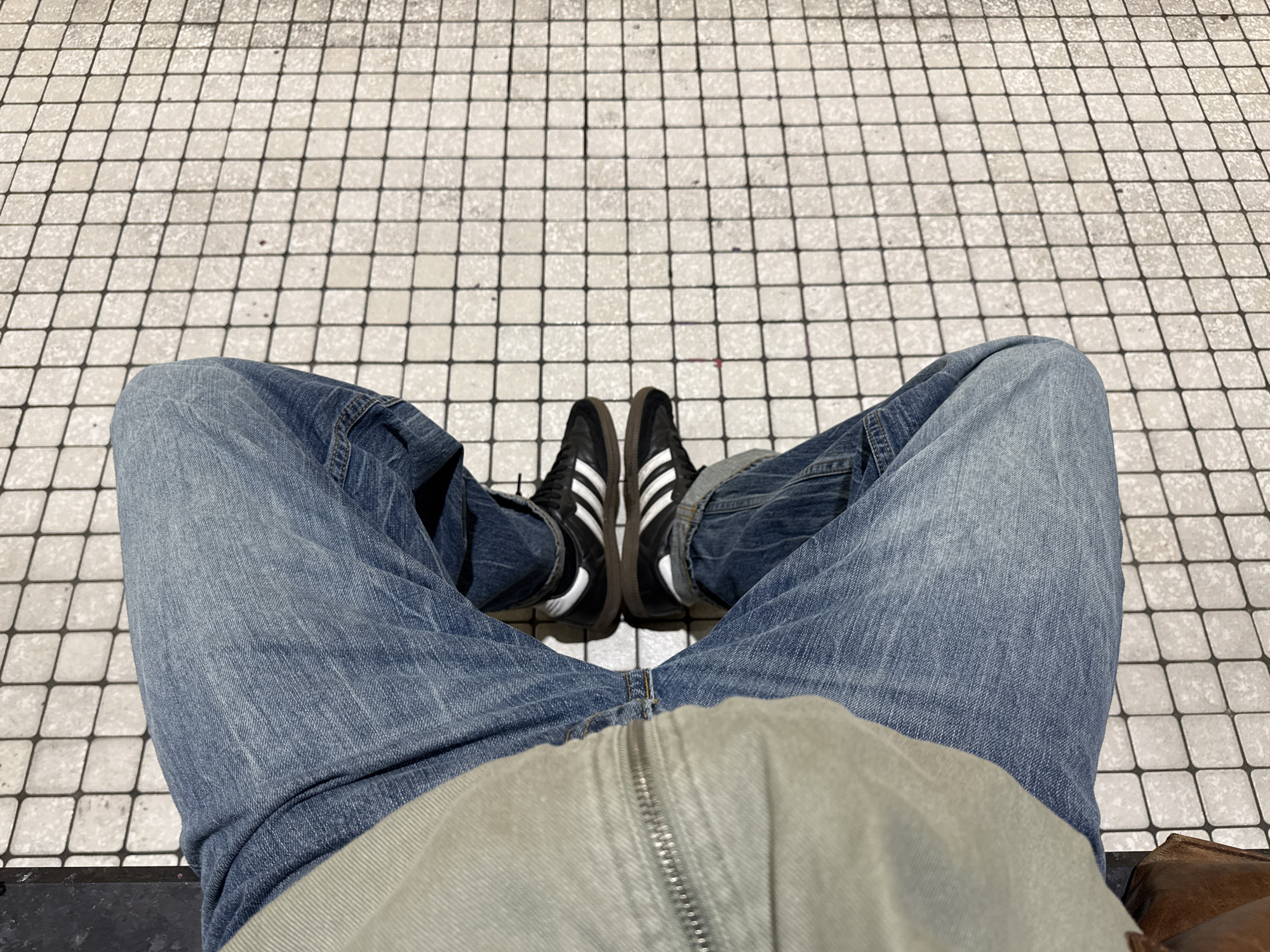 The image shows a first-person view of a person sitting, wearing blue jeans, black Adidas sneakers with white stripes, and a light gray jacket. The person is sitting on a surface covered in square tiles with black grout lines. The location is not discernible from the image provided.
