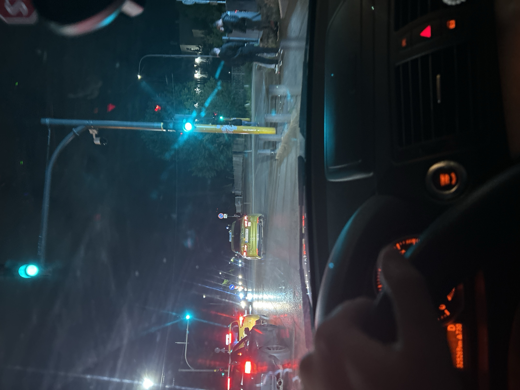 The image depicts a nighttime scene viewed from inside a car, likely in Sofia, Bulgaria. The car is stopped at a traffic light which is currently green. There are other vehicles in front, and pedestrians are visible on the sidewalk to the right. The dashboard of the car shows lit-up controls. The car's speedometer is illuminated. Streetlights and traffic lights are visible in the background, and the reflection of these lights is seen on the wet road surface, suggesting that it has recently rained.