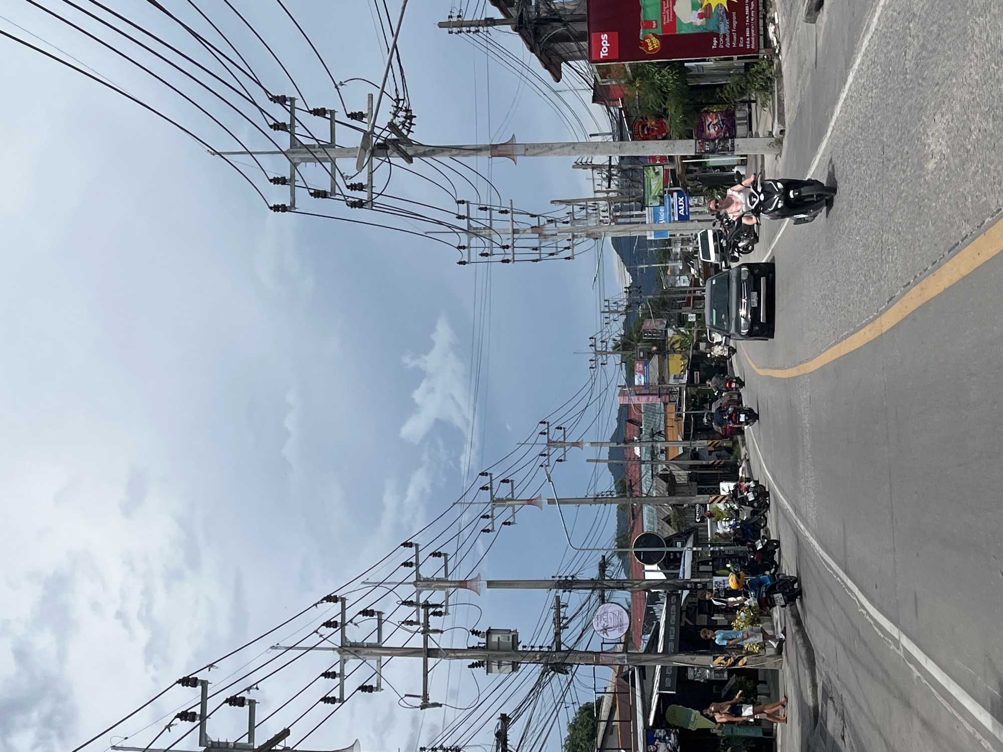 This media file captures a street scene in Tambon Ko Pha Ngan, Thailand, likely during the daytime under a cloudy sky. The image is dominated by a dense network of power lines and utility poles that stretch across the frame, creating a visual sense of overhead complexity.
Along the side of a paved road, motorbikes are parked and being ridden, indicating a busy thoroughfare. Several people are visible, some on motorbikes and others walking or standing on the sidewalk, which is lined with shops and businesses. One shop sign clearly reads "Tops" and another has the word "AUX" and "Midel". Another sign mentions "BAKERY & RESTAU" and shows what appears to be a product description for "Mixed Fragrant Rice".
The overall impression is one of a typical bustling street in a tropical urban or semi-urban environment. The presence of multiple motorbikes suggests a common mode of transportation in this region. The lighting suggests it's daytime, and the cloudy sky indicates potentially moderate weather.