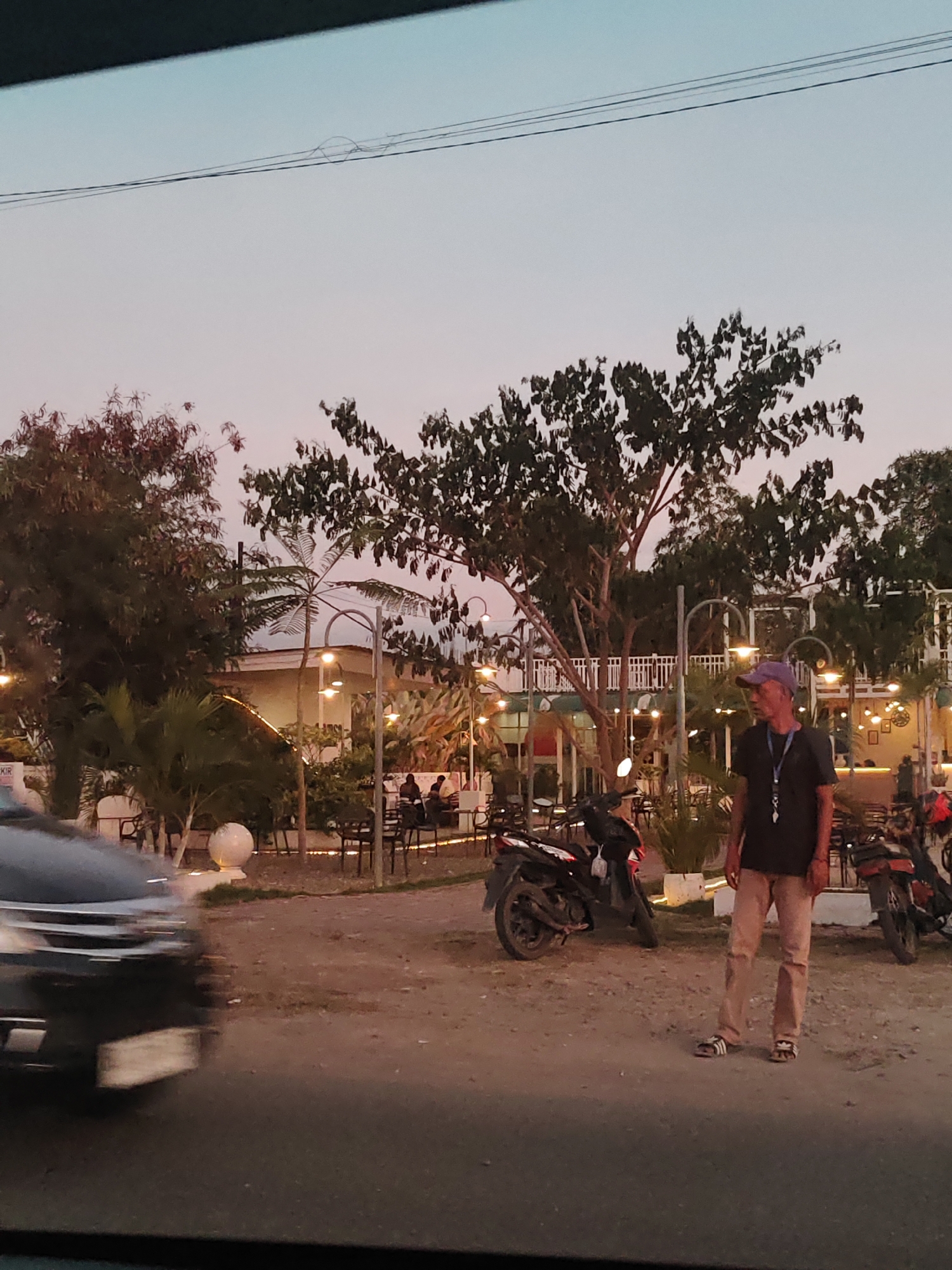 The scene shows a street in Cotabato, Indonesia, likely during the late afternoon or early evening due to the soft, dusky light. A car is partially visible in the lower left, blurring as if it is moving. A man stands on the right side of the image, wearing a purple baseball cap, a dark t-shirt with a lanyard around his neck, and tan pants. He wears open-toed sandals. Two motorcycles are parked near him, one to his left and another to his right. Behind the man, there is an outdoor dining area with lights, trees, plants, and furniture suggesting a restaurant or cafe. People are seated at tables. The ground appears to be dirt or gravel. Electrical wires run across the upper part of the sky.