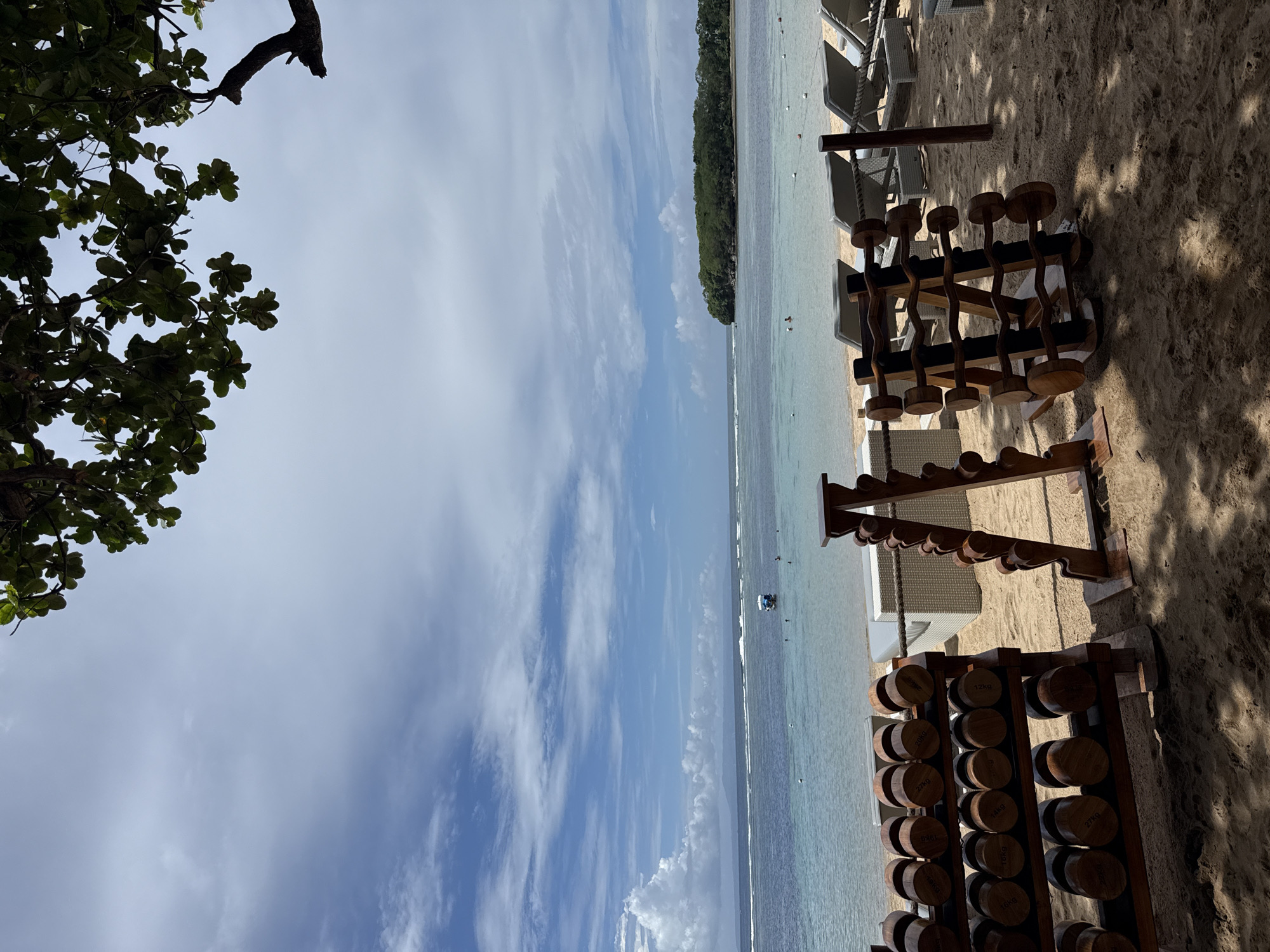 The image shows an outdoor exercise area on a beach, likely in Kuta Selatan, Indonesia, given the tropical setting. The foreground contains three racks of wooden dumbbells of varying weights. Behind the dumbbells, there are lounge chairs and a view of the ocean, which is clear and turquoise in color. A wooded area can be seen on the horizon, possibly an island or headland. The sky is partly cloudy with shades of blue visible through the clouds. Part of a tree and its leaves are present in the top-left corner of the image. The image is taken from a slightly elevated perspective.