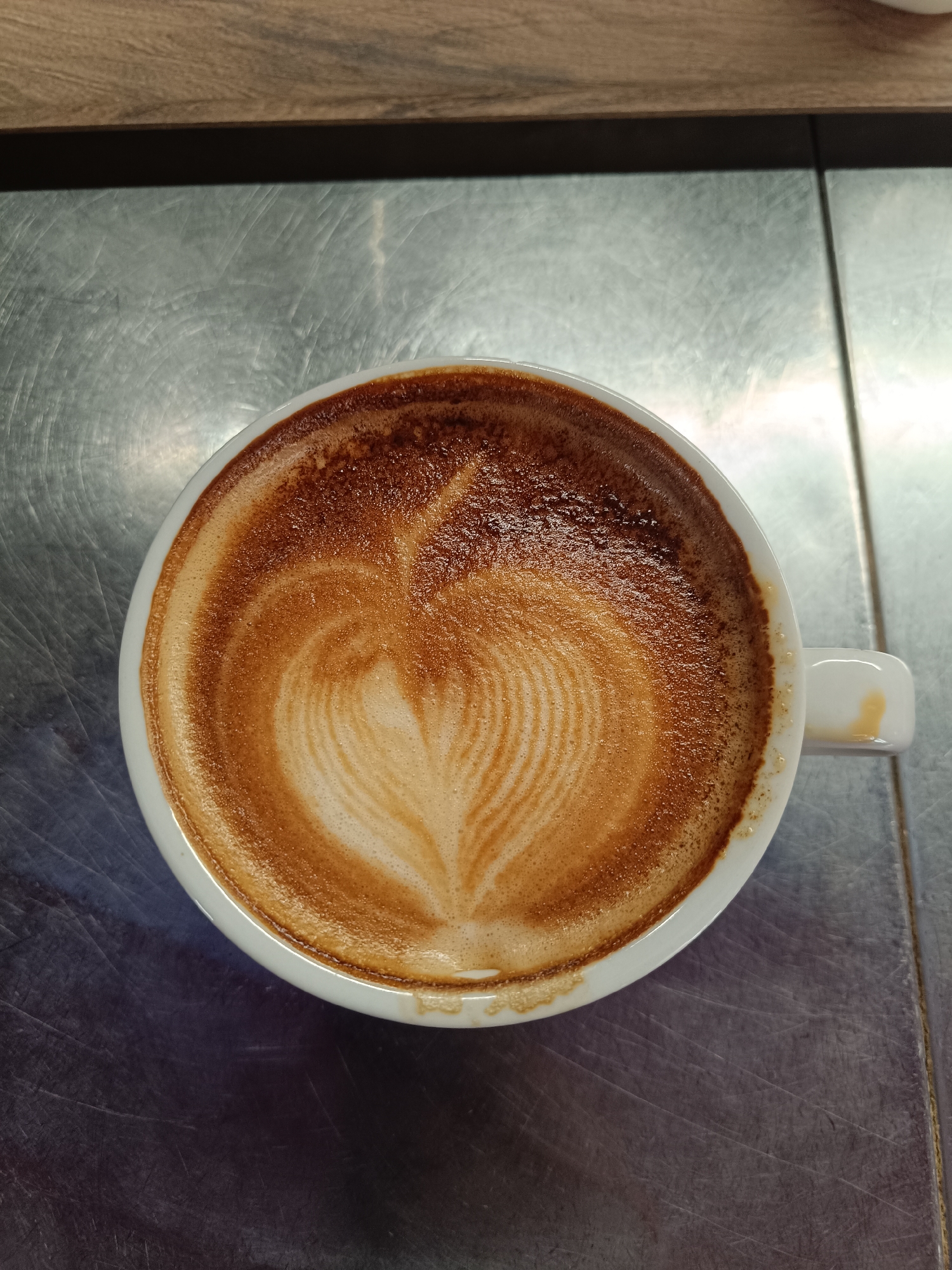 A top-down shot depicts a white ceramic coffee cup containing a latte with latte art. The art presents a heart-like pattern formed with concentric lines of coffee foam. The cup rests on a metallic surface, which shows signs of wear with visible scratches. The overall lighting is diffused, providing even illumination across the scene. There are no people visible in the image. It is impossible to ascertain the location in Plovdiv, Bulgaria, from the visual information alone.
