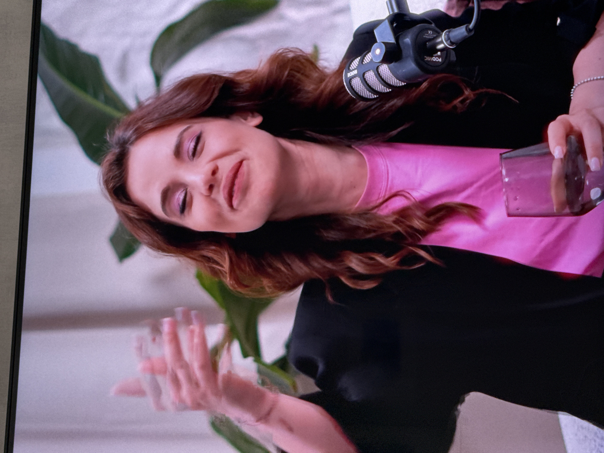 A fair-skinned woman with auburn hair is shown from the chest up, wearing a pink shirt and a black cardigan. She is holding a glass of liquid in her right hand. There is a microphone directly in front of her, and a potted plant with large green leaves is behind her to the left. The background appears to be a light-colored wall. There is no indication that the location is in Lovech, Bulgaria, or any specific city.