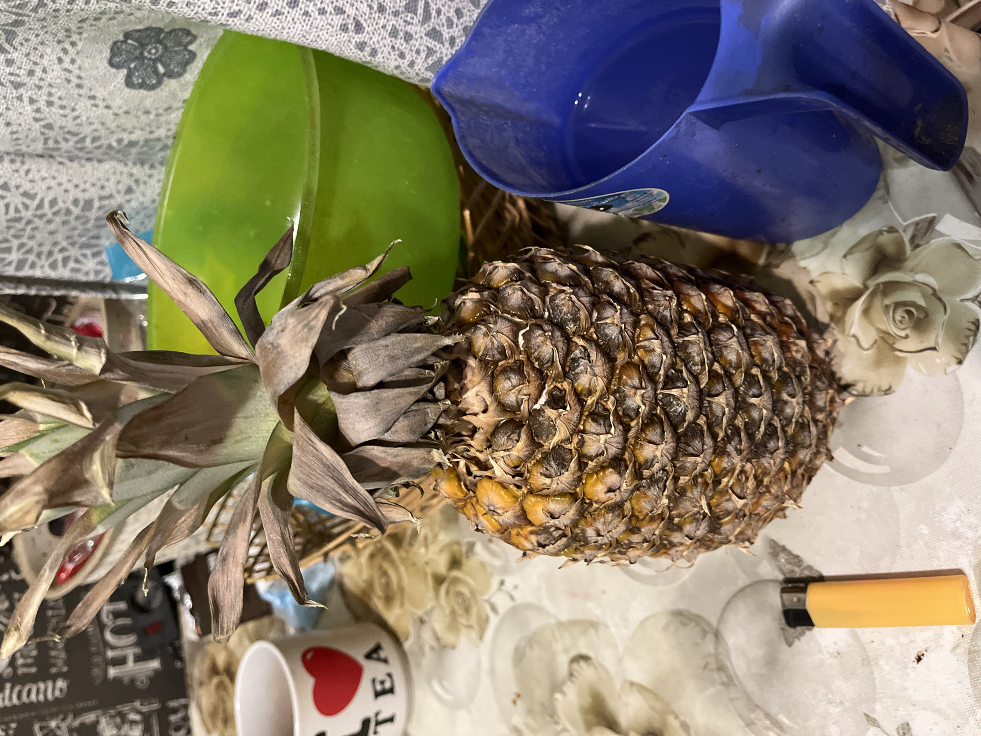 A pineapple is displayed on a table with a patterned tablecloth. To the left of the pineapple, a white mug with a red heart and the word "TEA" is visible. Behind the pineapple, a green bowl is partially submerged in a wicker basket. To the right of the pineapple, a blue pitcher contains liquid. An orange lighter lies on the tablecloth in the foreground. The scene appears to be indoors, likely in a domestic setting. There is no information within the image that suggests it is located in Minsk, Belarus.