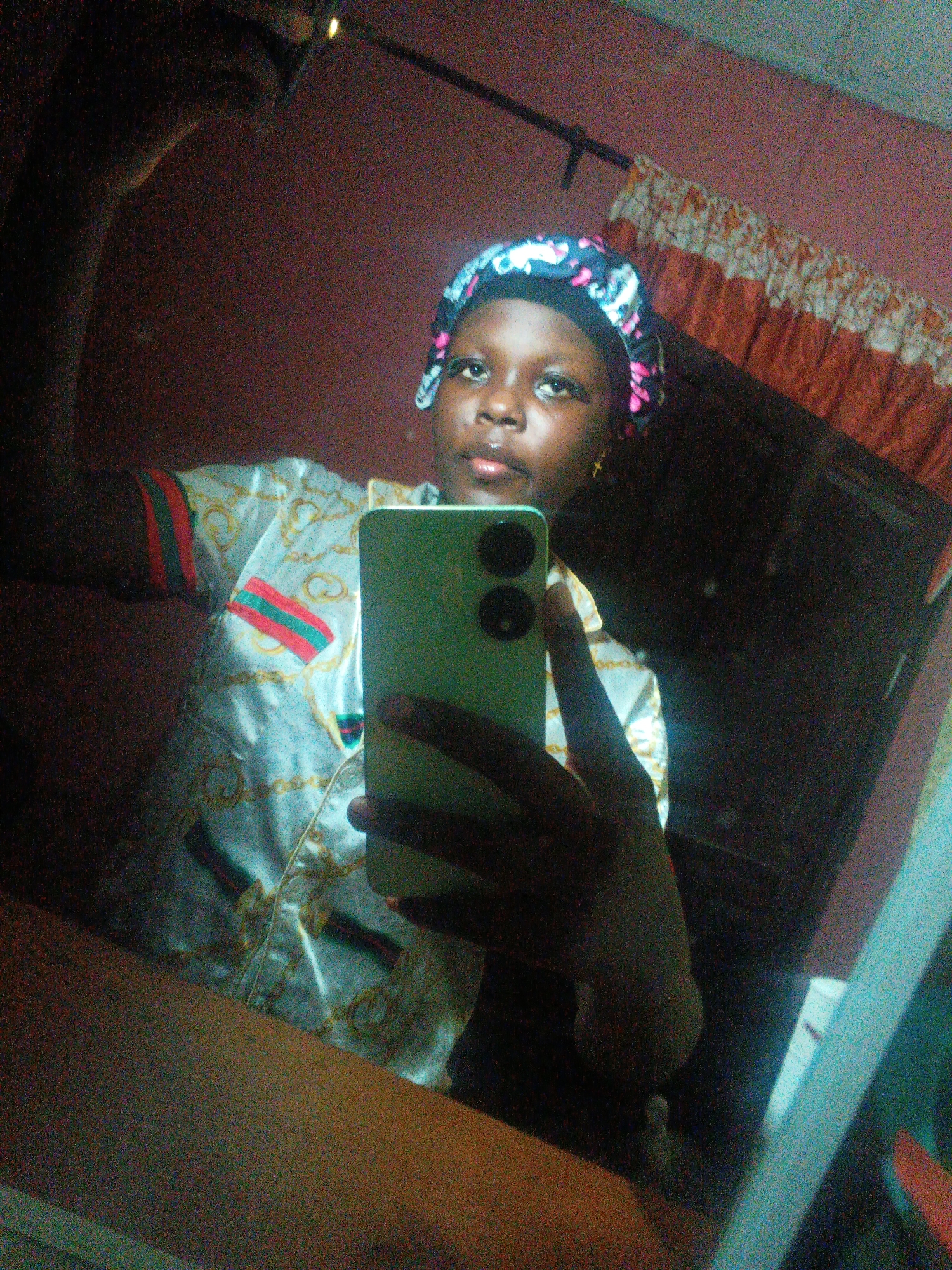 A mirror selfie captures a young person, likely a female, indoors in Onitsha, Nigeria. The subject, of African descent, is seen from the chest up, directly facing the camera with a neutral expression. She wears a colorful, patterned satin head covering, possibly a bonnet, and a light-colored short-sleeved shirt featuring green and red striped accents and gold chain-like designs. Small gold cross earrings are visible.
She is holding a vibrant green smartphone in her right hand, positioned in front of her lower body, obscuring part of her torso. The phone has two distinct camera lenses on its back. The image is a reflection, indicating the subject is taking a selfie in a mirror.
The background, visible in the mirror's reflection, consists primarily of a reddish-brown wall. In the upper right, a patterned curtain or fabric, featuring segments of orange and white/cream, hangs from a dark rod. The overall lighting appears somewhat dim, contributing to a noticeable level of digital noise throughout the image. There is no discernible text visible in the frame.