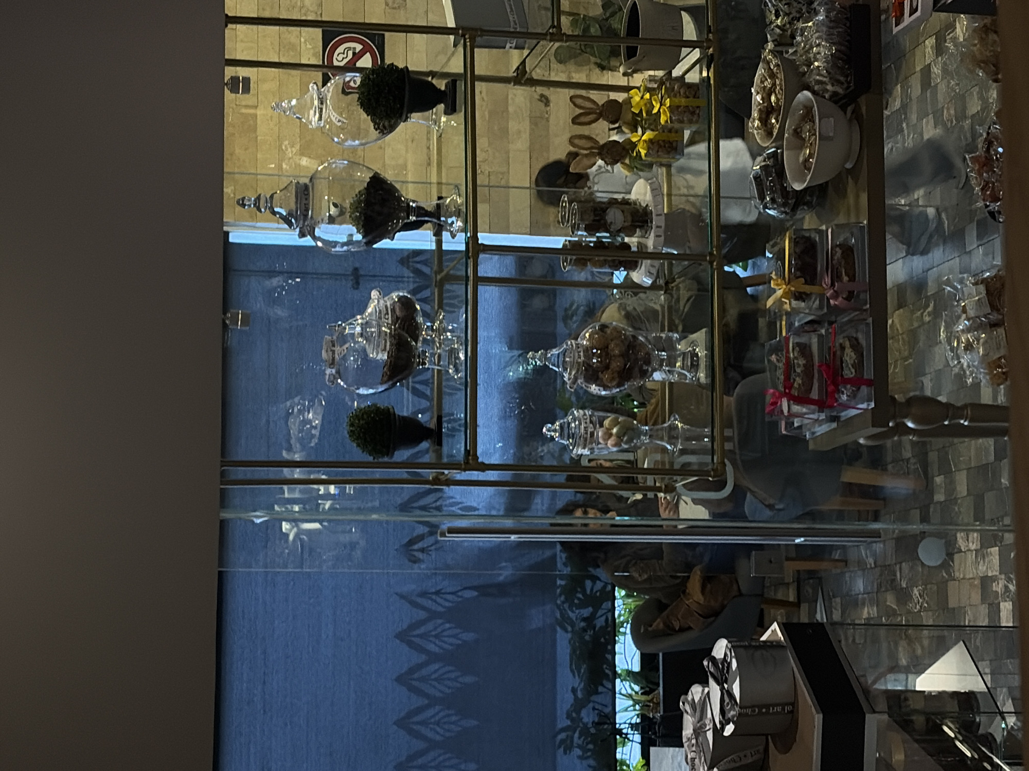 The image depicts the interior of a retail establishment, likely a confectionery or café, located in Puebla, Mexico. The scene is observed from a slightly elevated perspective, looking towards a display area and beyond into a seating section.
In the immediate foreground, a display table on the left holds two stacked cylindrical silver gift boxes with black ribbons. To the right, a lower glass shelf of a multi-tiered display unit is visible, showcasing various packaged items including small white bowls containing chocolates or candies, and several clear plastic boxes tied with red or yellow ribbons.
The central focus is a prominent three-tiered display unit, constructed from glass shelves supported by golden-toned metal uprights.
* The top shelf features three large, clear glass apothecary jars with lids. The leftmost jar contains dark granular material. The middle and right jars each hold a small, spherical green plant, resembling a topiary, situated in a black urn-like pot.
* The middle shelf displays a clear glass jar containing colorful oval candies, two cylindrical glass containers holding cookies or similar items, and two decorative brown rabbit figures with yellow accents, accompanied by small yellow-ribboned gift boxes. A white pot is also present.
* The bottom shelf largely mirrors the lower foreground, displaying more packaged products in bowls and clear boxes.
Through the clear glass partitions and doorway of the display unit, the background reveals a seating area. To the left, a person with dark hair, wearing a dark top, is seated in a dark gray armchair, with a brown jacket or bag draped over the chair. This individual holds a white cup. Another person is partially obscured further back. Behind the seating area, a large window or glass wall is covered by a dark blue roller blind with a repeating light-colored leaf pattern along its lower edge. Green foliage is visible beyond this window. To the far right, an exterior wall with light-colored brick or stone texture features a circular red and white "No Smoking" sign. The floor of the establishment is covered with square tiles in varying shades of gray. The ceiling is dark, with visible recessed lighting fixtures.