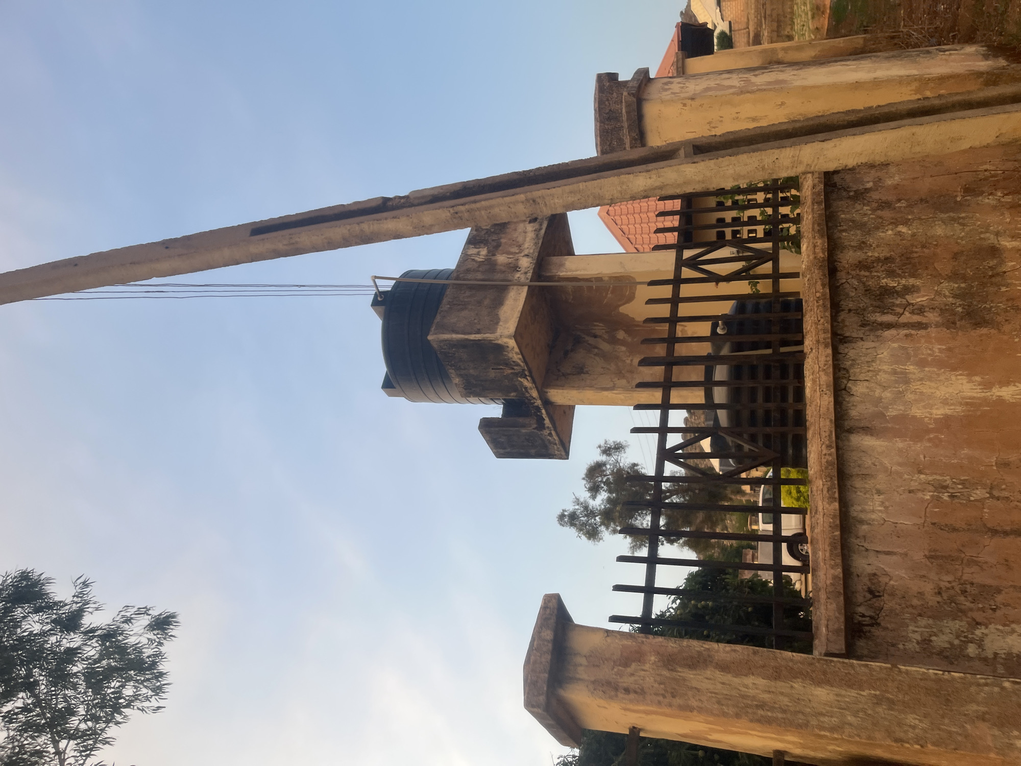 The image depicts an outdoor scene in Barakin Akawo, Nigeria. A large, black water tank sits atop a concrete structure, with a long, angled concrete beam extending towards the sky. A weathered concrete wall with a black metal fence runs along the right side of the frame, partially obscuring a building with a red-tiled roof in the background. Trees are visible through the fence. The sky is light blue.