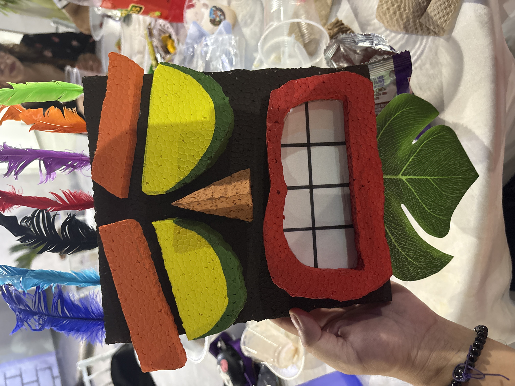 A craft project resembling a tiki mask is held in a person's left hand, in a close-up, eye-level shot. The mask is primarily black with colorful foam and feather accents. The mask features prominent orange foam eyebrows above large yellow circular eyes outlined in green foam. A small brown triangular nose is centrally located. The mouth is wide and red, with white foam teeth forming a grid pattern. A green leaf shape is attached to the bottom of the mask. Several colorful feathers, including blue, light blue, black, red, purple, orange, and green, protrude from the top of the mask. The background is blurred and shows a white tablecloth with various objects scattered on it, including plastic cups, wrappers, and a bottle of red liquid. In the far background, out of focus, a person wearing glasses and a black patterned shirt is seated at a table. The setting appears to be an indoor event or gathering. There is no visible indication of the city of Progreso, Mexico within the image itself.