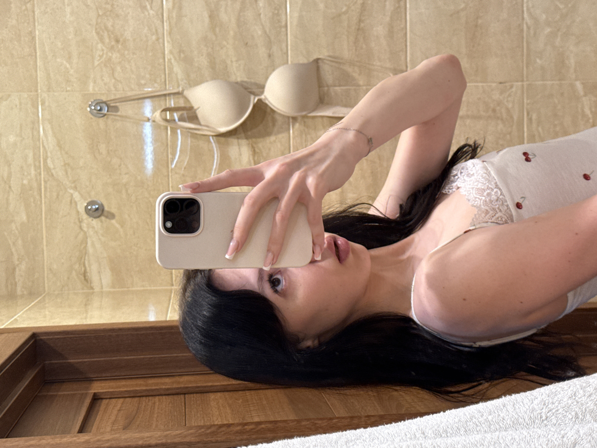 A young woman with long, dark hair is taking a mirror selfie in a bathroom. She is wearing a light-colored tank top with lace trim and cherry print. She is holding a phone in her right hand to take the photo, obscuring her face. In the background, a beige bra is hanging on a hook on a tiled wall. A wooden door is visible on the left side of the image, and a white towel is hanging next to it. The scene appears to be in a private residence. The city of Khimki, Russia is mentioned in the prompt but there are no visual cues in the image to confirm this location.