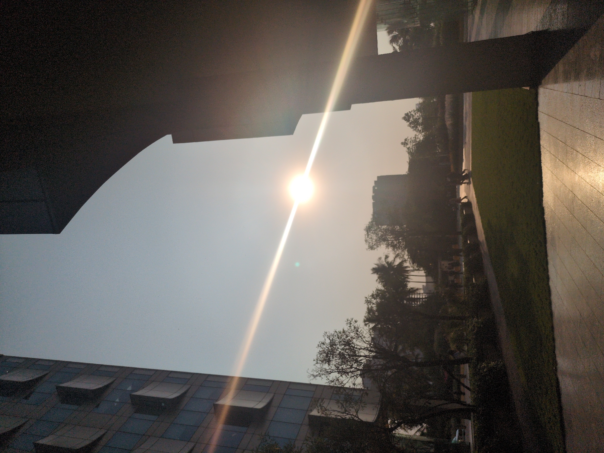 The image shows a low-angle view towards the sky in Bengaluru, India. The sun is bright, creating a strong glare. Buildings are visible at the bottom and in the background, one with multiple rectangular window sections. Trees are present, partially obscuring the view. Part of a dark structure, potentially a roof or awning, is visible in the upper left corner of the frame. A walkway with shiny, possibly wet, tiles borders a green grassy area on the right. The lighting suggests it may be morning or late afternoon. Some people can be vaguely detected walking on the opposite sidewalk, near the tree line.