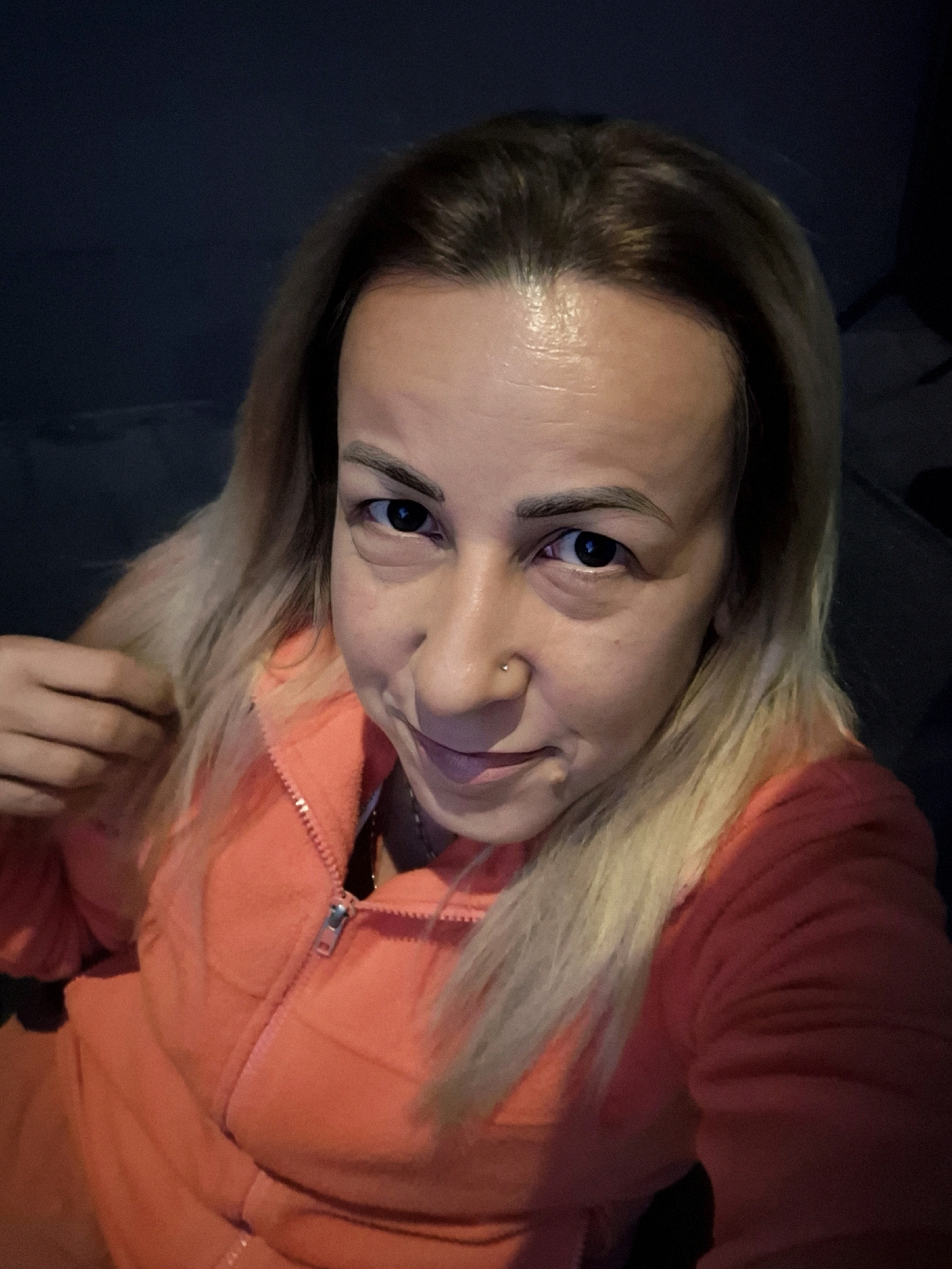 A selfie is captured from a high angle, looking down at a Caucasian woman with blonde hair. She is wearing an orange zip-up hoodie. Her hair is light blonde and extends to her shoulders. She has a small silver nose ring. The background is dark and out of focus, suggesting an indoor setting at night. The woman's skin appears to be slightly oily. She is touching her hair with her left hand. Her mouth is slightly open in a smile.