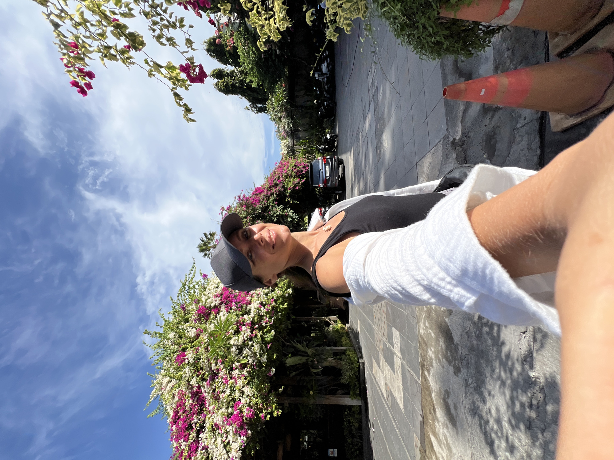 The image depicts a selfie taken by a woman wearing a black tank top, a white shirt draped over her shoulders, and a grey baseball cap. She has a warm smile and is looking towards the camera. The background features lush vegetation with vibrant pink and white bougainvillea flowers cascading over a walkway. A dark-colored SUV is parked behind her, and further in the distance, several motorcycles are visible, suggesting an outdoor parking area. The ground is paved with grey tiles. The sky above is a clear blue with wispy white clouds. The scene appears to be an outdoor area within a tropical or subtropical city, likely a public space or the grounds of a hotel or resort. The presence of a car and motorcycles, along with paved walkways and manicured plants, indicates an urban or semi-urban environment. Given the flora and overall ambiance, the location could be Denpasar, Indonesia, a city known for its tropical climate and vibrant natural beauty.