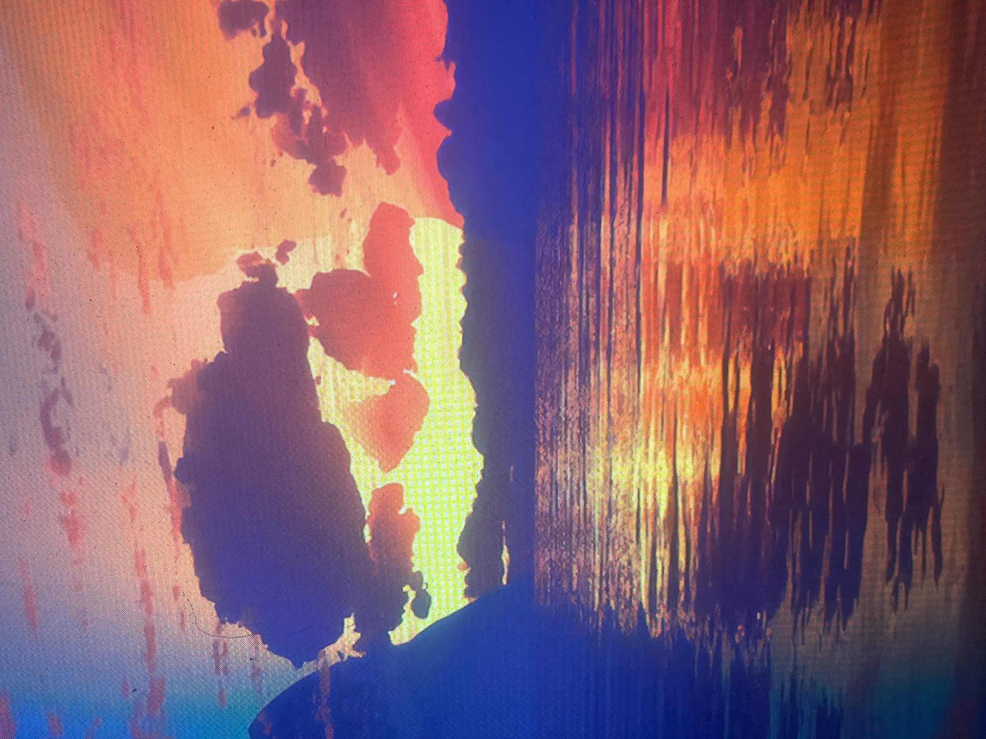 The image displays a highly saturated, pixelated landscape, appearing to be a photograph captured of a digital screen rather than a direct photograph of a natural scene. The depicted location is cited as Zelenyi Hai, Ukraine, though no specific geographical markers within the image confirm this attribution.
The scene features a prominent body of water in the foreground, reflecting an intensely colored sunset or sunrise sky. The sky transitions dramatically from a deep blue on the far left, through purples and oranges, to vibrant reds on the right. Large, dark, amorphous cloud formations are silhouetted against the bright sky, with a powerful yellow-orange light source, presumably the sun, emanating from behind a central cloud mass just above the distant horizon. Smaller, scattered reddish clouds are also visible.
A dark, sharply peaked landmass is present on the far left, rising above the water. Along the horizon, beyond the expanse of water, are additional dark, irregular silhouettes of distant land or mountains. The water's surface reflects the vivid sky and the bright light source in elongated, horizontal, wavy streaks of blue, purple, yellow, orange, and red.
No individuals, animals, or man-made structures are present within the scene. The entirety of the image, including all crops, exhibits a distinct diagonal grid pattern and visible pixelation, consistent with the moiré effect and screen door artifacts from photographing an illuminated digital display. Several small dust specks are also visible on the image surface, particularly in the upper left quadrant.