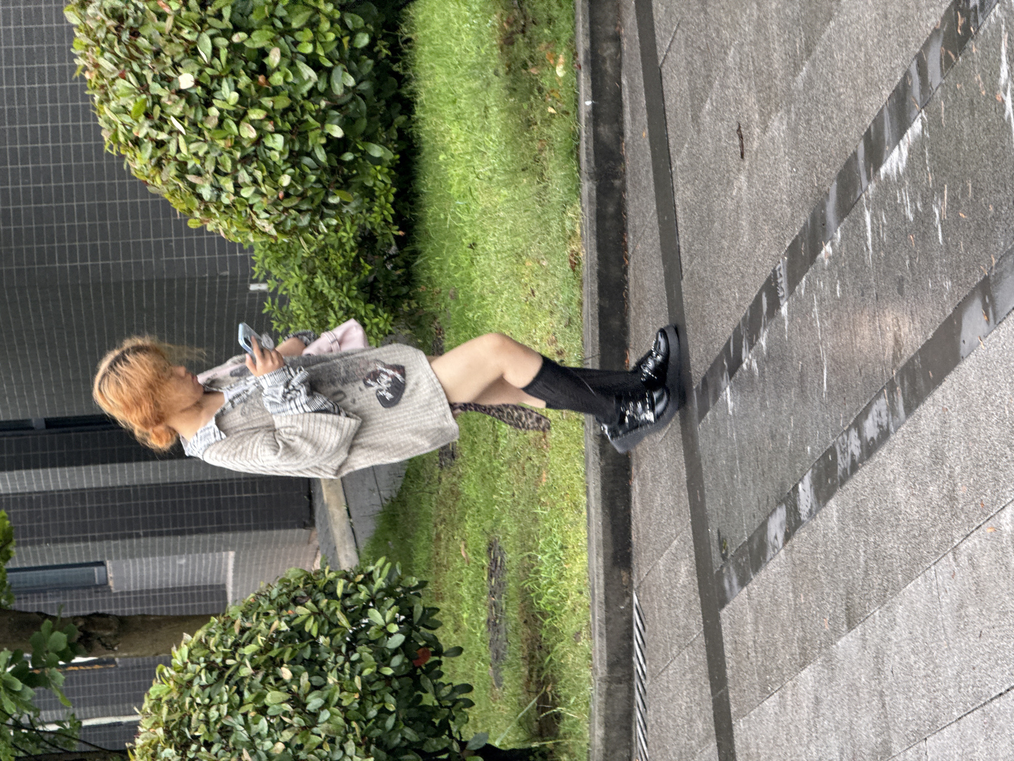 A sideways, medium-angle, outdoor shot shows a fair-skinned person with blonde hair standing on the sidewalk in Jiangjin, China. The person is angled slightly to the left, facing away from the camera, and wearing a patterned, light gray sweater, black thigh-high socks, and black shiny shoes. The individual is holding a phone to their face.
The sidewalk is gray concrete with darker gray bands. Green grass lines the sidewalk, with bushes on both sides. A gray and dark gray wall is behind the individual.