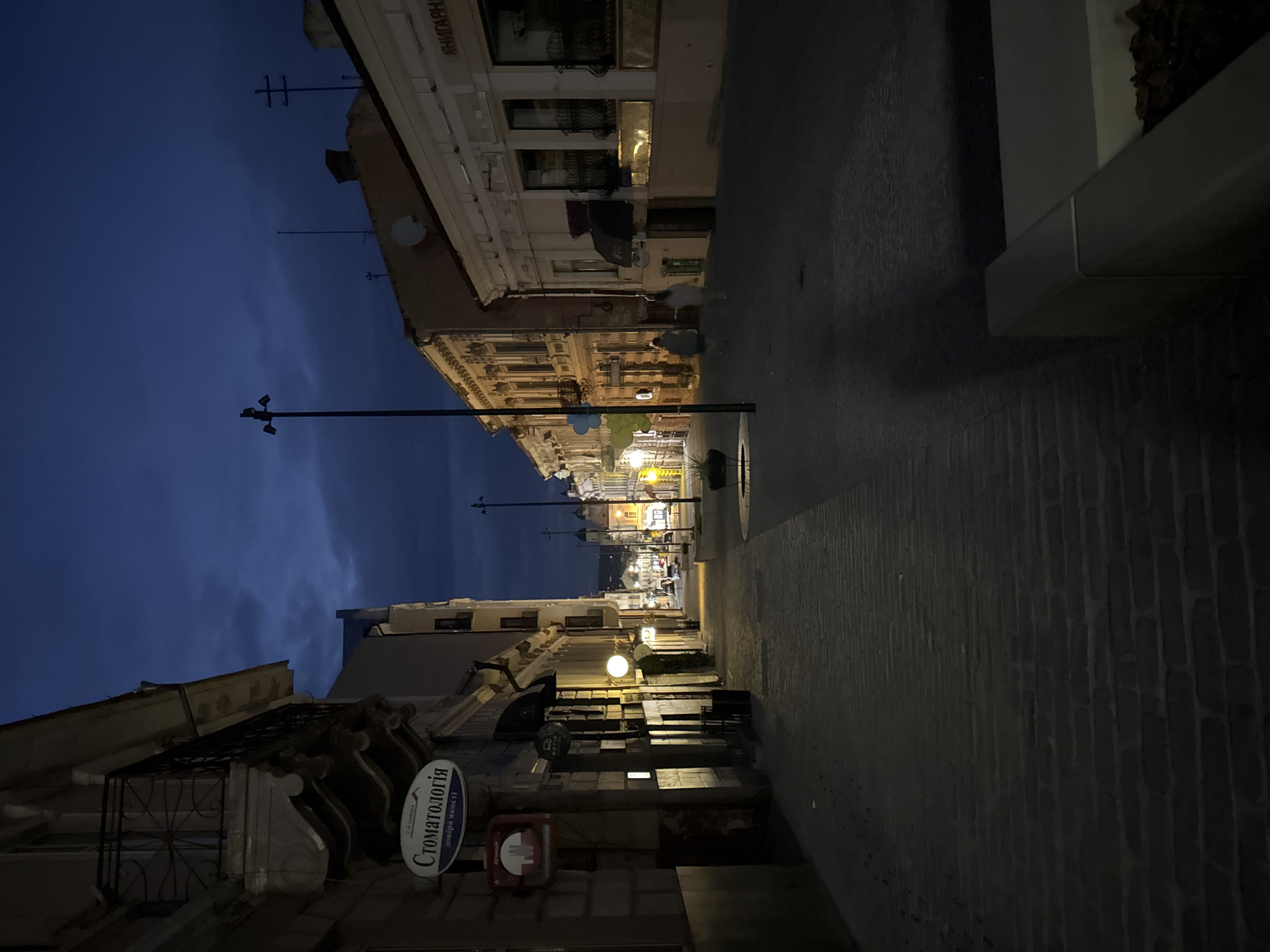 The image depicts a street scene at dusk or night in Chernivtsi, Ukraine. The street is paved with cobblestones and is lined with buildings of various architectural styles, some featuring ornate facades and balconies. Streetlights illuminate the pathway, casting long shadows and highlighting the details of the architecture. A few people are visible walking along the street, their forms softened by the dim lighting. Signs for businesses, including a "Stomatolohiya" (Dentistry) sign, are visible on the buildings. The sky is a deep blue, indicating late evening or early morning, with some cloud cover. The overall atmosphere is quiet and serene, with the urban environment illuminated by artificial light against the darkening sky.