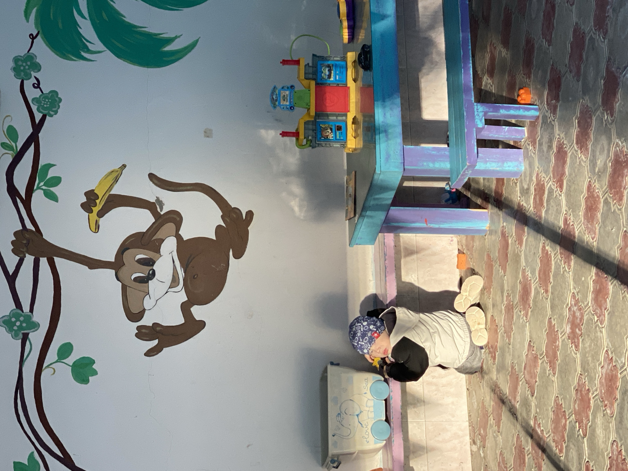 This media file captures an indoor scene, likely a playroom or a daycare, featuring a child and various toys against a vibrant wall mural and a tiled floor.
The main subject is a young child, possibly a toddler, who is seen from above and slightly behind, kneeling or sitting on the patterned floor. The child wears a blue patterned beanie hat, a light grey or white long-sleeved top over a dark shirt, and light-colored sneakers. They appear to be engaged in play, holding a small yellow object in their hands near a white toy container.
The room's left wall (when oriented correctly) is adorned with a colorful mural depicting a brown cartoon monkey holding a yellow banana, swinging from a brown vine with green leaves and light green flowers. Above the monkey, large green palm-like leaves are painted. Visible cracks run vertically on the white wall.
On the floor, near the child, is a white toy container with light blue wheels and an elephant design. The words "love" and "Sea" are visible on its side. To the right, a multi-colored toy structure, predominantly yellow, red, and blue, with what appears to be character images in blue panels, sits on a blue and purple wooden-like play table or structure. An orange toy lies on the floor further to the right.
The floor is covered in distinct grey/brown tiles with a repeating reddish-orange floral or geometric pattern. Strong shadows are cast across the floor and parts of the structures, indicating bright lighting, likely from a window or a strong overhead light source. The overall impression is a cheerful and child-friendly environment.