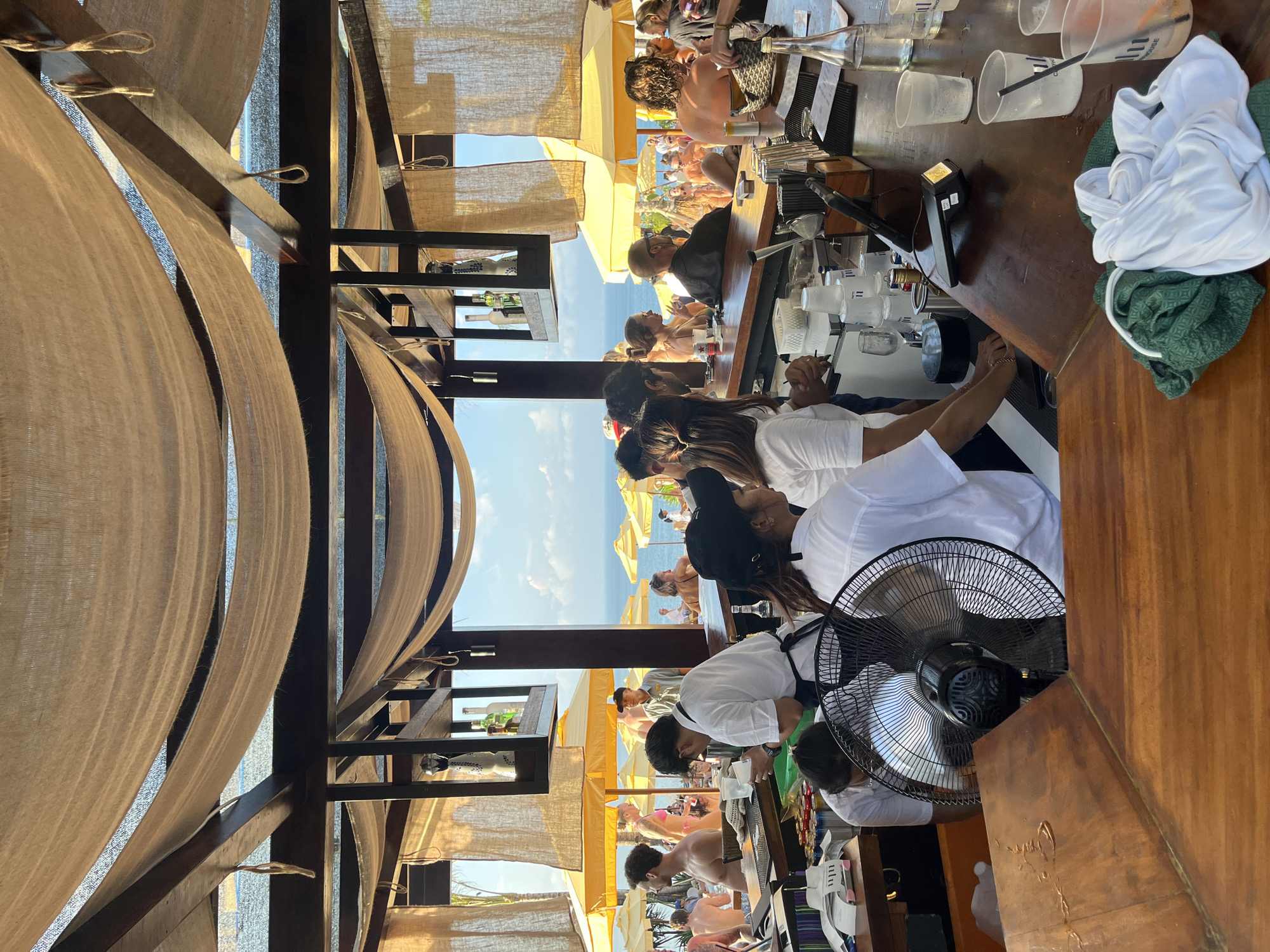 The image depicts an outdoor bar in Kuta Selatan, Indonesia, likely a beach club setting. Several bartenders are working behind a wooden bar counter. Customers are seated at the bar and beachgoers can be seen in the background, under yellow umbrellas. The bar is covered by a beige canvas-draped structure. Visible on the bar counter are various glasses, bar tools, a point-of-sale device, and folded clothing. A black electric fan is positioned near the front of the counter. The ocean and sky are visible between the structure and beach patrons.
